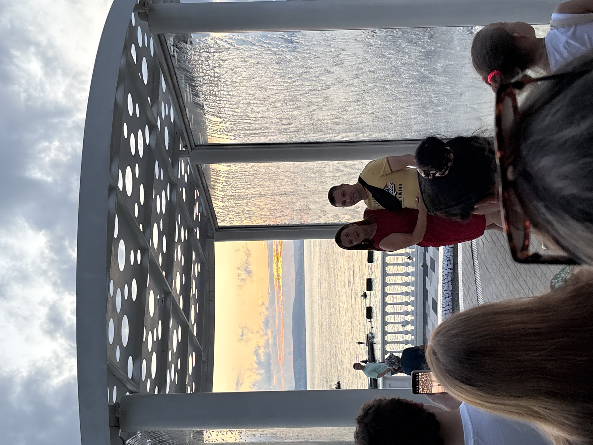 This image depicts a couple posing for a photo in front of a waterfall feature, likely in Novorossiysk, Russia, given the sunset over the sea. The couple is standing under a white, circular-patterned canopy, with water flowing down transparent panels on either side of them. The woman is wearing a red dress, and the man is in a yellow t-shirt and black fanny pack. In the background, a picturesque sunset paints the sky with warm hues over the sea, with a distant cityscape visible on the horizon. Several people are in the foreground, some observing the couple and others taking pictures with their phones. The overall atmosphere is one of leisure and tourism, possibly at an outdoor venue or promenade along the Black Sea coast.