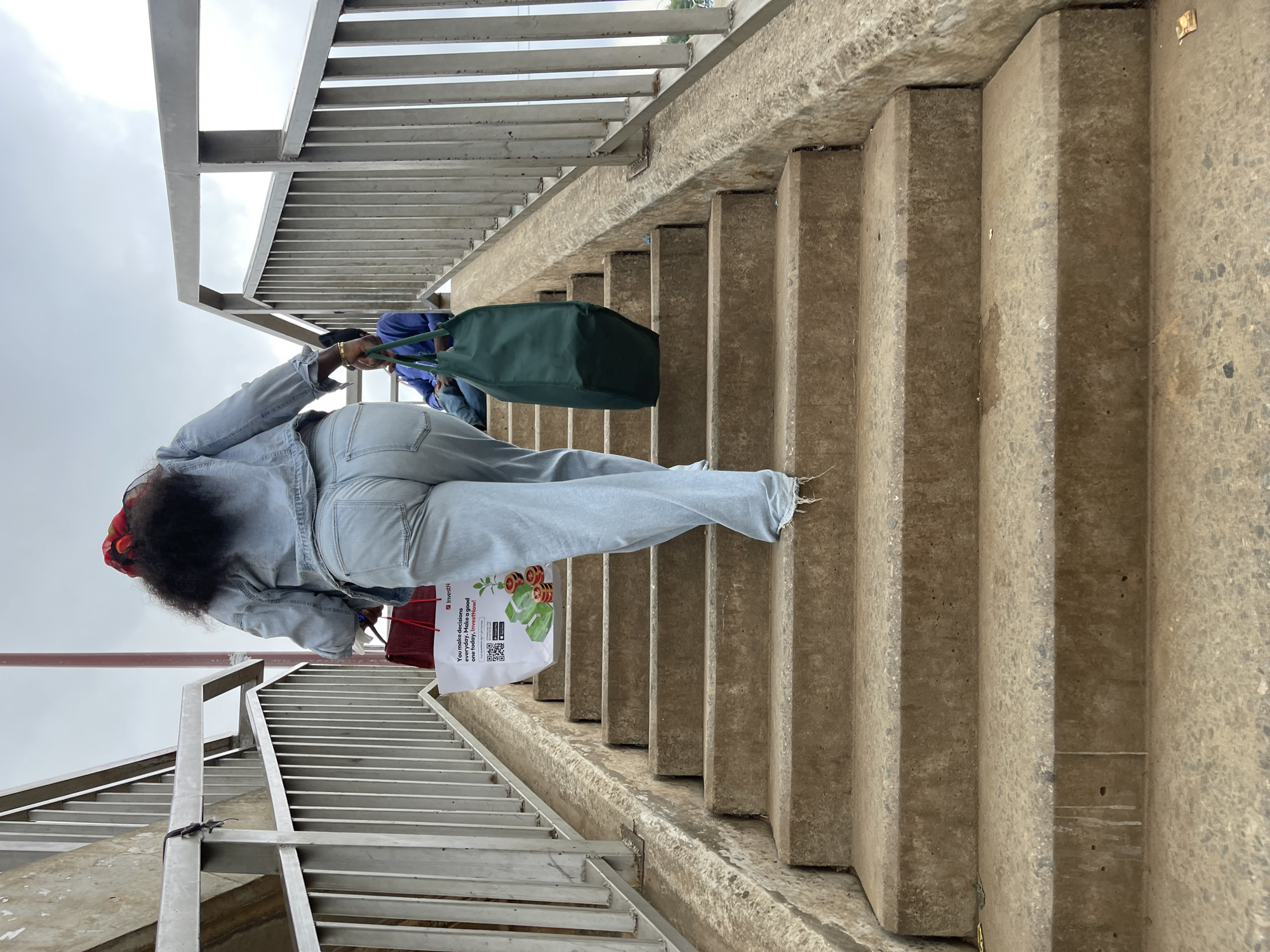 The image depicts a person walking up a concrete staircase, likely part of a pedestrian overpass in Gwarinpa, Nigeria, based on possible location clues. The person, viewed from behind, is wearing light blue denim jeans and a matching jacket. They are carrying multiple bags, including a green tote bag and a white bag with the word "InvestN" and an advertisement printed on it. The staircase has metal railings on both sides. The sky is visible in the background.