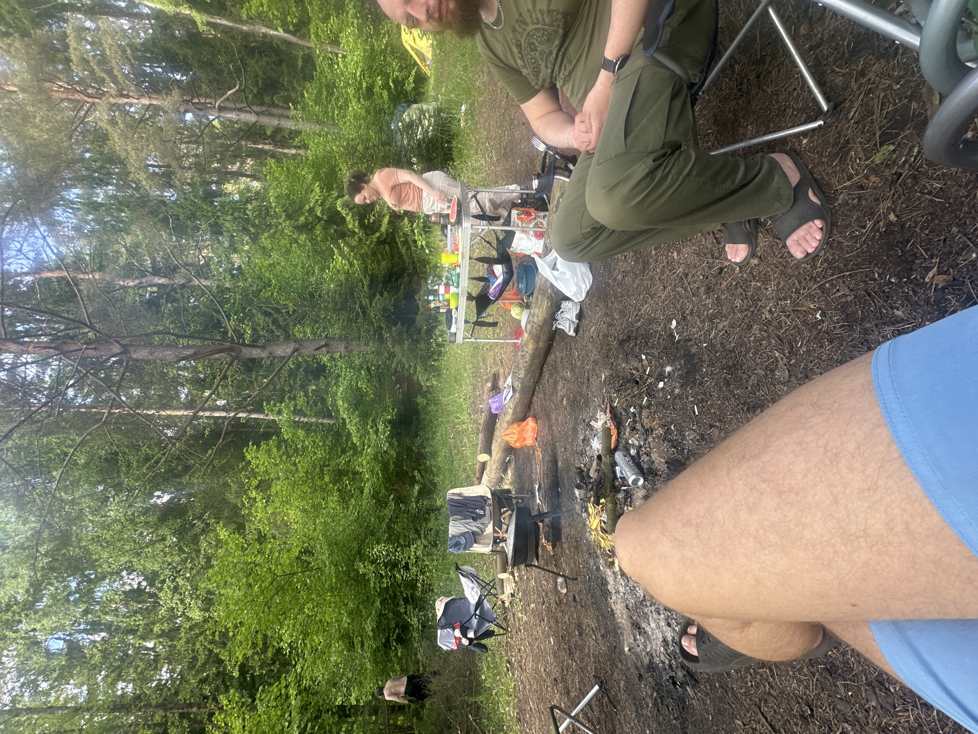 The image depicts a campsite scene in a wooded area, potentially within the Юзуфоўскі сельскі Савет region of Belarus. A woman stands near a folding table laden with food and supplies. A man with a beard is seated in a folding chair. Another person wearing blue shorts is seated near a campfire. A metal tripod cooking stand is positioned over the fire. Trees surround the campsite. Tents are partially visible in the background.