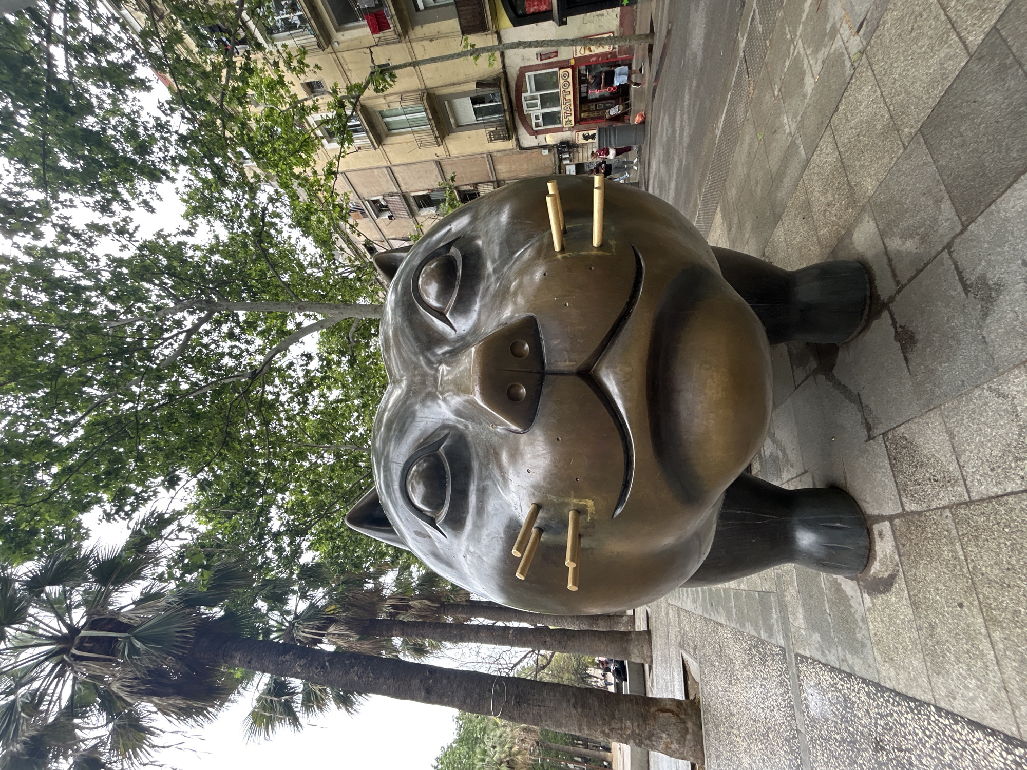 The image shows a close-up of the "El Gato de Botero" sculpture, a bronze cat by Colombian artist Fernando Botero, located in Barcelona, Spain. The sculpture is positioned on a pedestrian walkway composed of gray and beige tiles. The cat is large and appears squat, with an oversized head and stout limbs. Its facial features are exaggerated, featuring large eyes and whiskers made of golden rods.
In the background, blurred buildings with multiple floors and balconies are visible, along with trees of various sizes. Some people are discernable in the distance, suggesting a public space. There is also a shop with "TATTOO" written on it. The lighting suggests an overcast day.