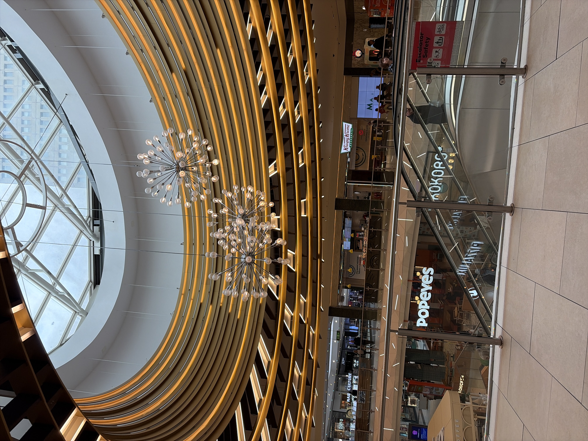 The image captures the interior of a multi-story shopping center in Manchester, United Kingdom. The building features an open, circular design with a glass ceiling that allows natural light to flood the space. Below the ceiling are decorative light fixtures resembling exploding stars. Each floor is lined with various shops and restaurants including Krispy Kreme, Popeye's, and Kokoro. Escalators are visible behind glass barriers, alongside signs warning about escalator safety. There are reflections of people on the glass walls.