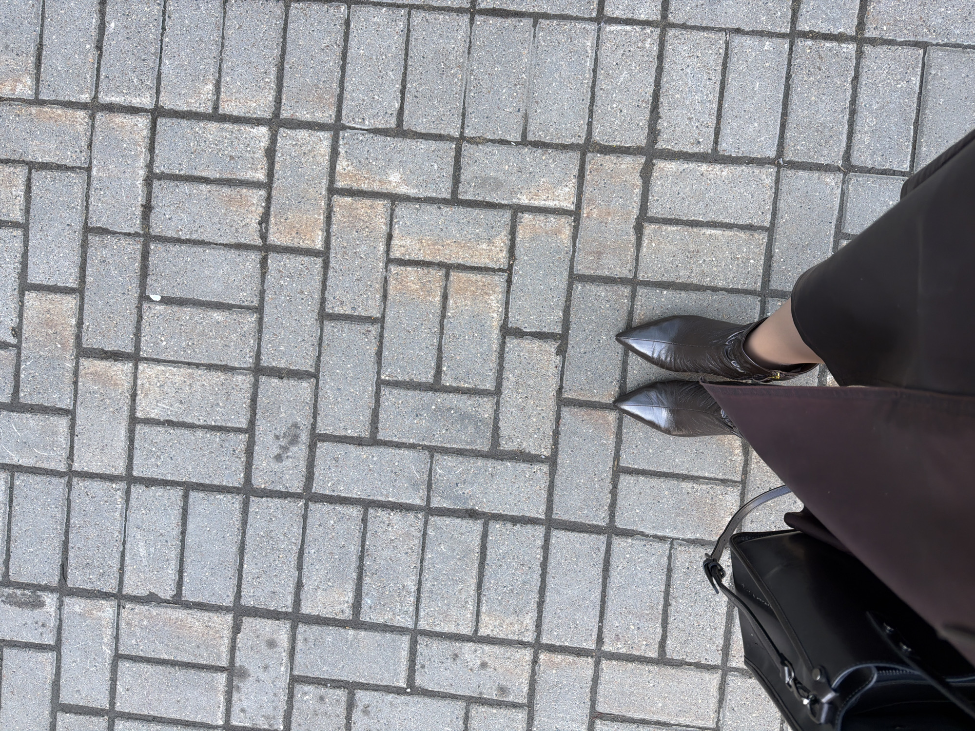 The image shows a person standing on a paved surface made of rectangular gray bricks. The person is wearing brown pointy-toed ankle boots and a brown skirt or dress. A black handbag is also visible. The location is implied to be Izhevsk, Russia.