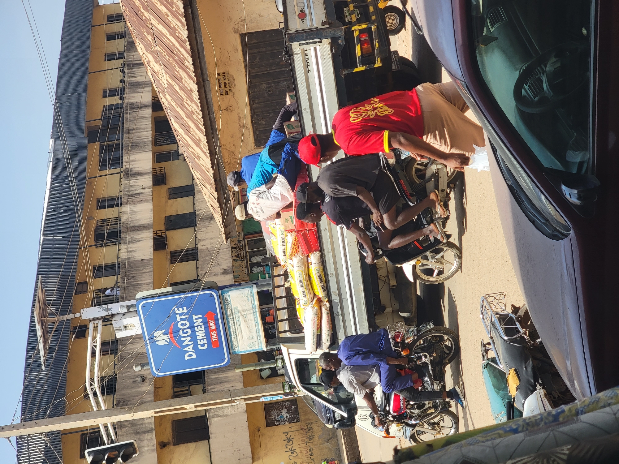 In Maiburiji, Nigeria, a medium-angle daytime shot shows multiple people gathered around a flatbed truck. Several men are loading bags labeled "MR. RICE" and other goods onto motorcycles. The men are dressed in a mix of casual clothing, including red, blue, and black shirts, and some wear hats. One person is visible inside the flatbed. Parked motorcycles line the street near the truck. A "DANGOTE CEMENT" sign is visible on the building above a traffic light. The street is lined with buildings of varying heights and architectural styles. A maroon car is partially visible on the right side of the frame. Street-level graffiti is evident on the buildings. The angle of the photograph is slightly tilted.