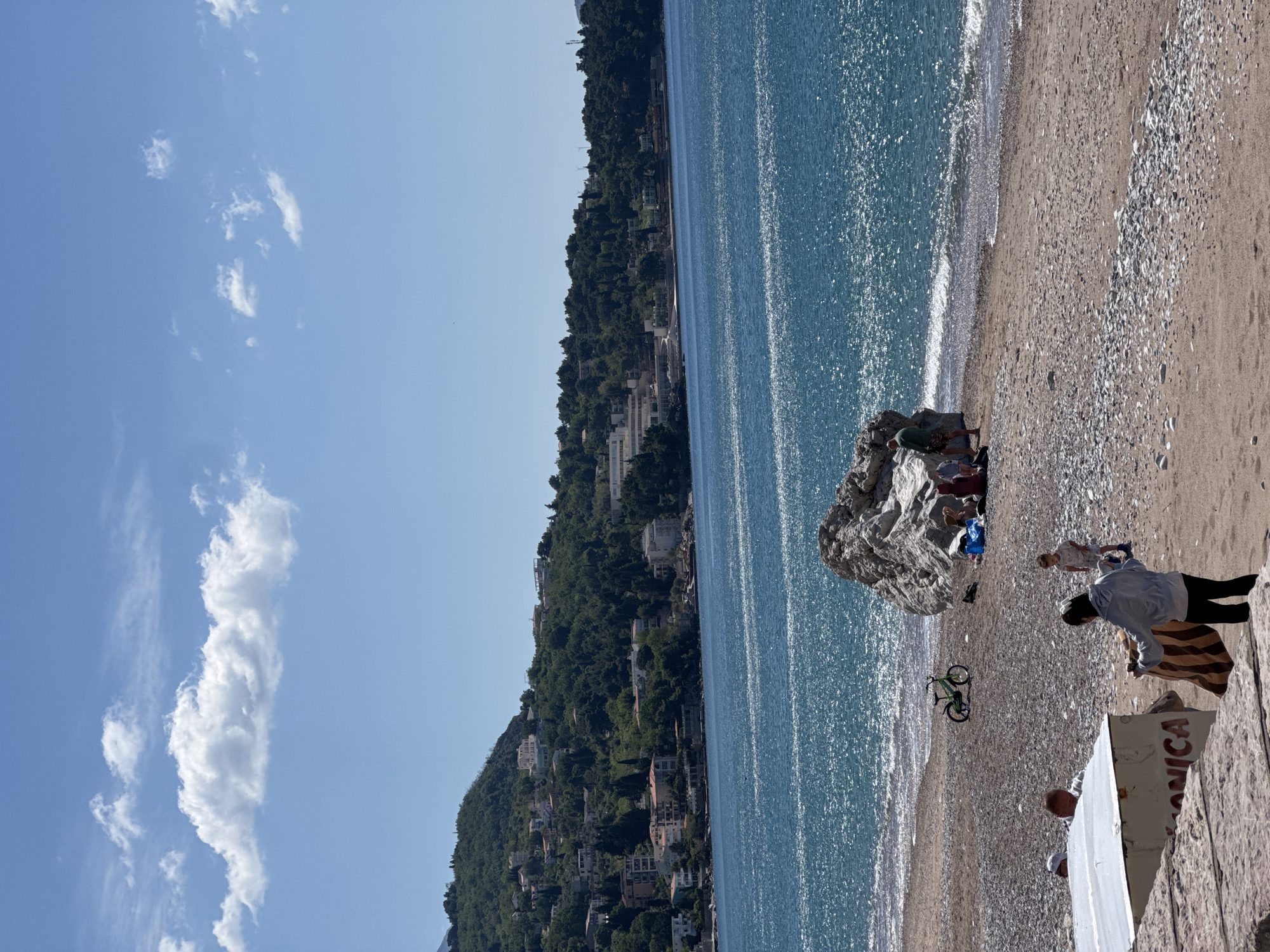 This image captures a vibrant coastal scene in Zankovići, Montenegro, under a clear, bright sky. The original image and its crops are oriented sideways, but assuming an upright view:
The upper portion of the frame features a bright blue sky dotted with scattered white cumulus clouds, indicating fair weather. Below, the sparkling blue waters of the Adriatic Sea stretch across the middle, reflecting sunlight with numerous glittering points. Gentle waves can be seen lapping onto the shore.
To the left, a steep, green hillside covered in dense trees rises from the coastline. Numerous buildings, likely homes or hotels, are nestled amongst the foliage, characteristic of a Mediterranean coastal settlement.
The foreground reveals a beach composed of both sand and pebbles. On the beach, a large, light-colored rock stands prominently near the water's edge. Several people are gathered around and behind this rock, appearing to relax. Further to the right, a person in a light blue shirt and dark pants stands beside a striped blanket on the sand. A green bicycle is parked upright near the water. In the bottom right corner, a white boat is partially visible, with the word "ANICA" clearly painted in red letters on its side. Portions of other individuals can be seen near the boat.
The scene depicts a sunny and peaceful daytime setting, showcasing the natural beauty of the Montenegrin coast combined with human leisure activities.