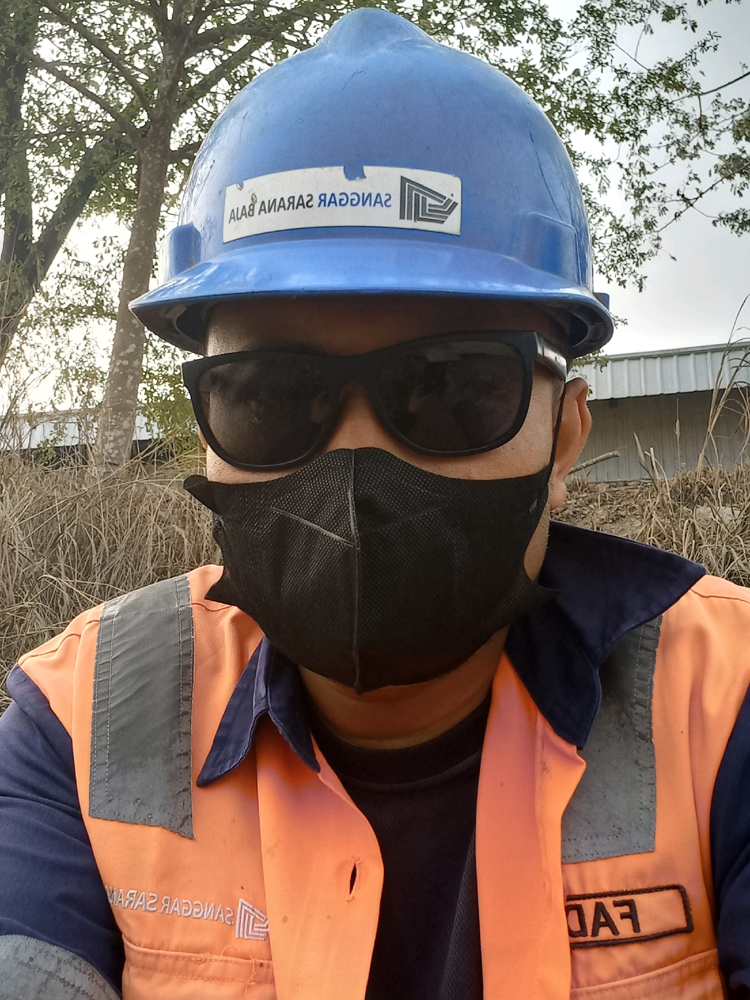 A person wearing a blue hard hat, black sunglasses, a black face mask, and an orange safety vest over a navy blue shirt is pictured outdoors. The hard hat has a white sticker with a logo and text that reads "ALAB AMAЯAZ ЯAOOZUA". The vest has reflective gray strips on the shoulders and a black name tag with white text that reads "FAЯ". The person is standing in front of dry grass and trees, with a metal-roofed building in the background. The image appears to be a selfie taken by the subject. The text on the hard hat and vest appears to be in Indonesian, suggesting the photo was taken in Indonesia. Based on the reflective strips and the type of building, this location could be an industrial area.