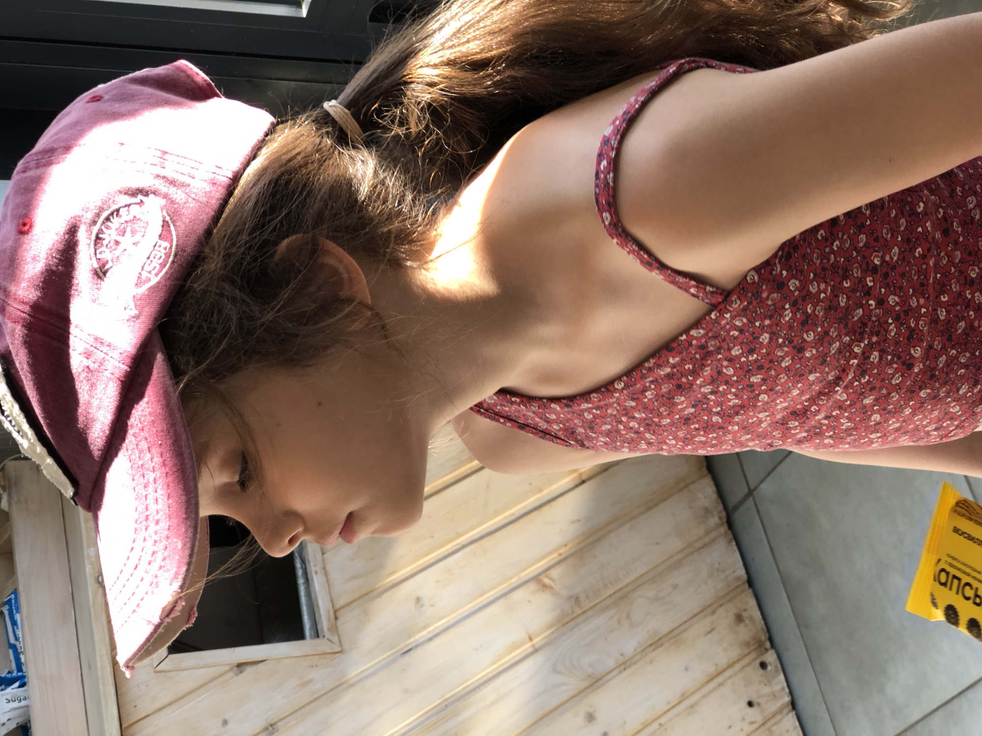 The image shows a young girl in profile wearing a red baseball cap and a red floral dress. She is looking to the left with her eyes closed. Her brown hair is pulled back in a ponytail. The background consists of a wooden wall and a tiled floor. A yellow wrapper with black writing is visible on the floor in the lower left corner. This image does not provide any information about the location in the city of Saint Petersburg, Russia.