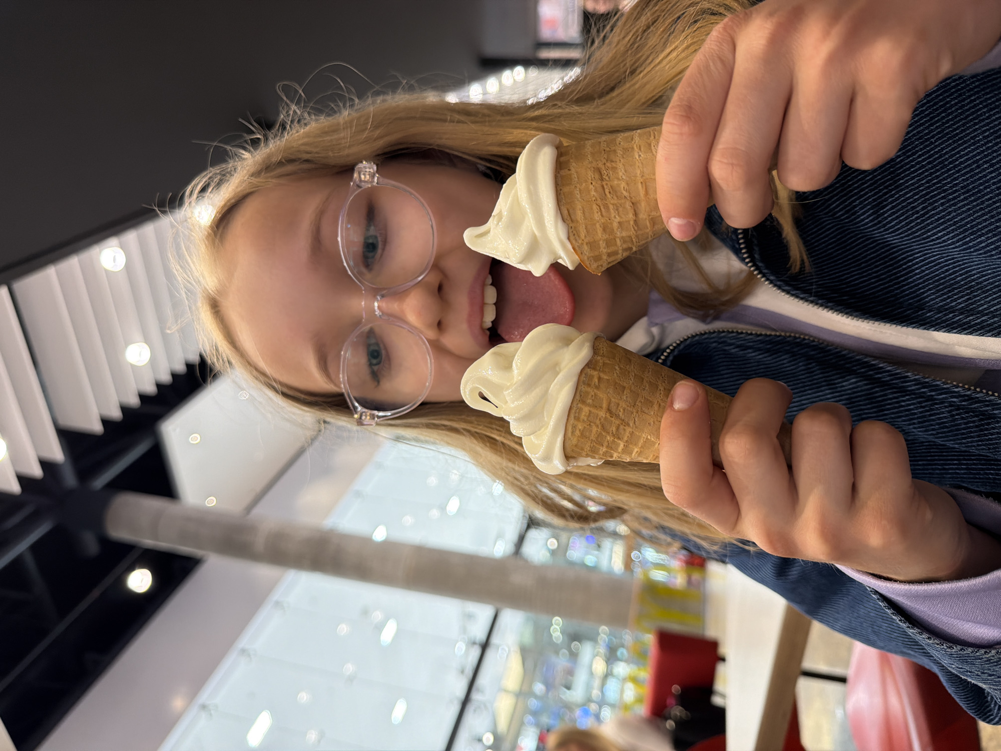 A young, blonde-haired Caucasian girl with glasses is holding two soft-serve ice cream cones. She is wearing a blue denim jacket and a light purple top. The girl is sticking her tongue out towards one of the ice cream cones. The background suggests an interior space, possibly a shopping mall, given the visible lighting fixtures, store displays and other people. The location is in Riga, Latvia.