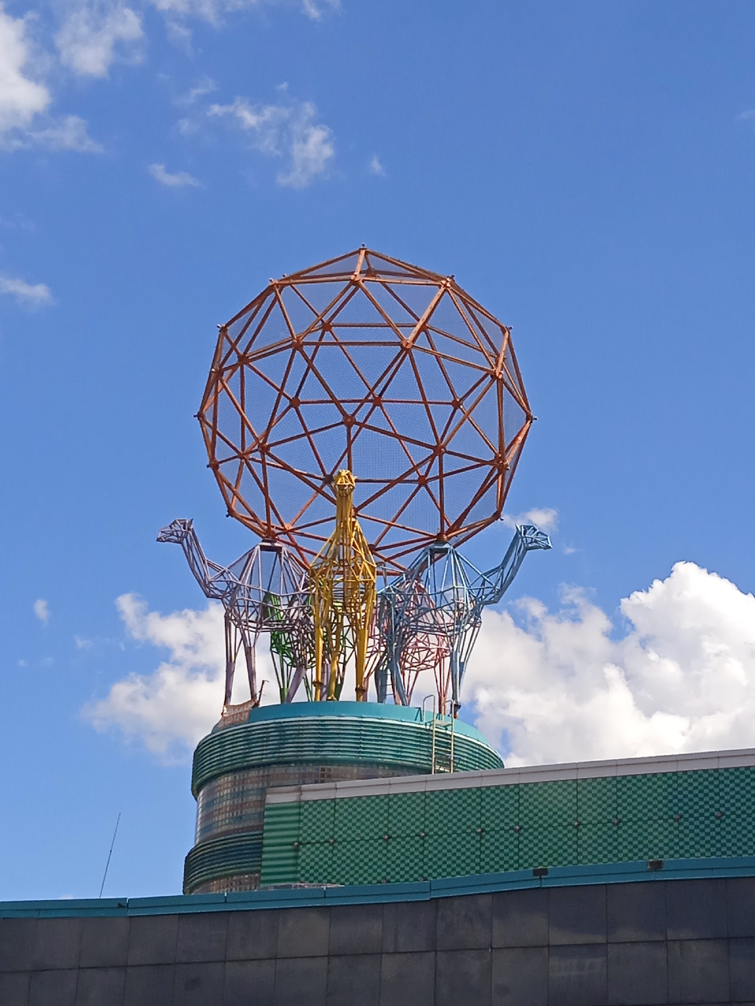 A geodesic dome-like sculpture, made of orange metal, sits atop a building in a city. The sculpture is supported by wireframe animal figures, including camels and what appears to be a giraffe. The animals are constructed from colorful metal wire, with some appearing in purple, blue, yellow, and green. The building beneath the sculpture has a rounded, tiered structure with horizontal green bands and a checkered green facade. The sky above is a bright blue with scattered white clouds.