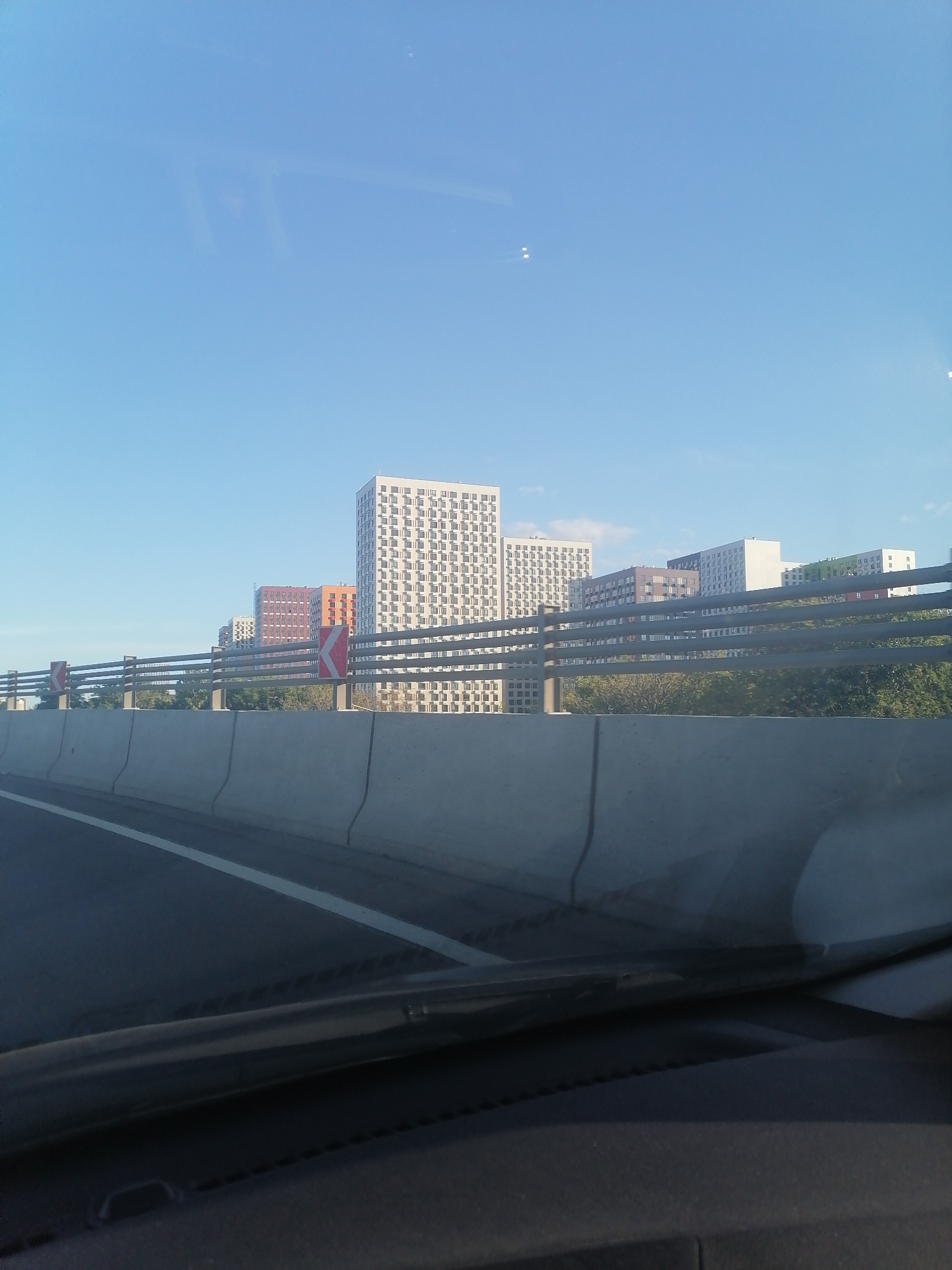 This is a photograph taken from a vehicle on a clear day, showing a highway with a concrete barrier and guardrail. Beyond the highway, several modern residential buildings are visible, including a tall, white building with many windows. The buildings are arranged in a row, with some colored in orange, red, and green. Trees and greenery are present between the highway and the buildings. The sky is clear blue. The scene appears to be in a suburban or urban area of Moscow, Russia.