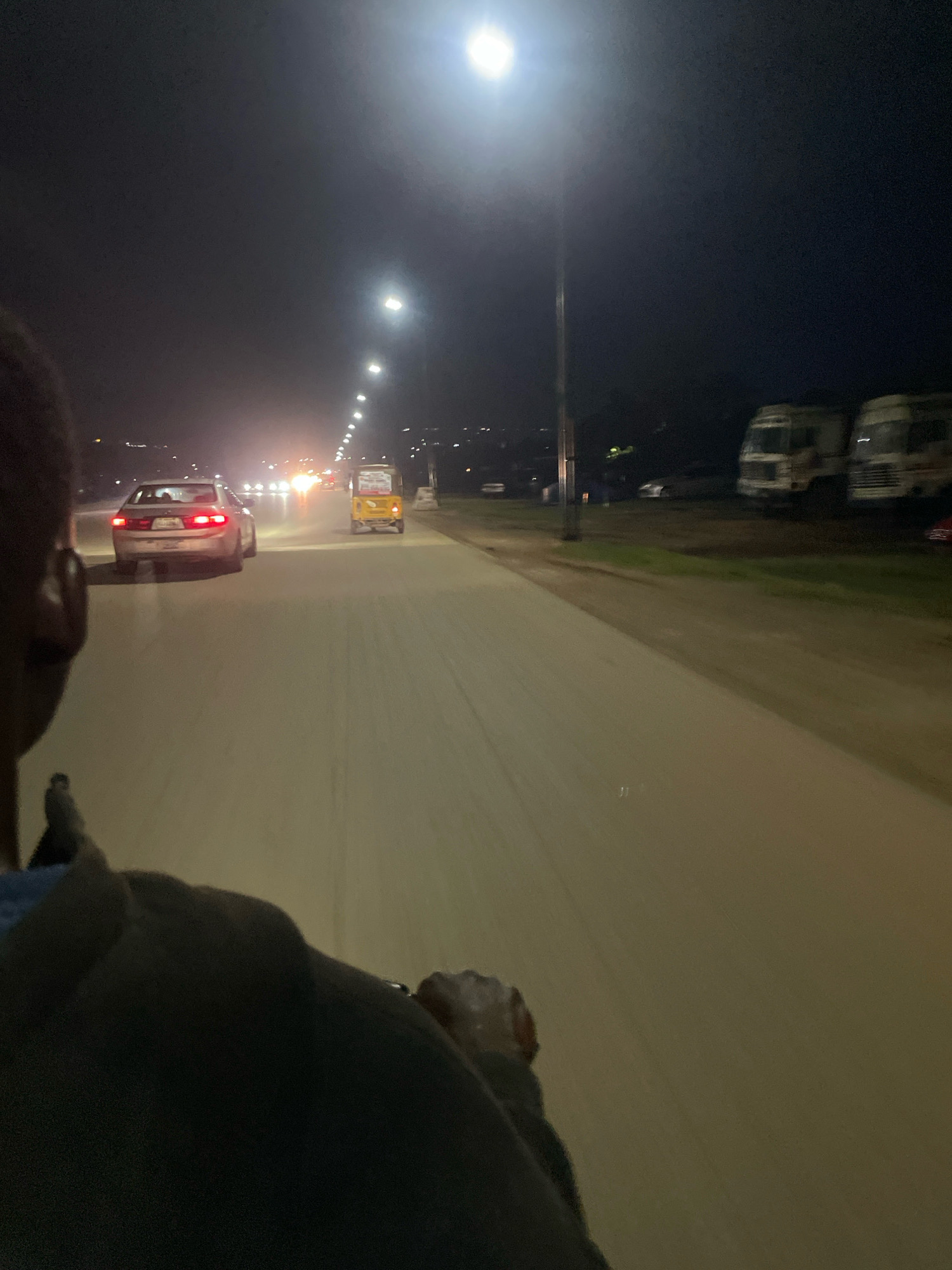 The image captures a scene at night on a paved road in Abuja, Nigeria, likely a highway or major thoroughfare given the presence of streetlights and multiple lanes of traffic. The perspective is from a vehicle, possibly a motorcycle or tricycle, looking forward. In the foreground, the back of a person's head and shoulder are visible, wearing a dark garment.
Further ahead on the road, a silver sedan is traveling in the same direction, its brake lights illuminated. To the right of the sedan, a yellow auto-rickshaw, also known as a Keke Napep in Nigeria, is visible. The road is well-lit by streetlights, creating a path of light stretching into the distance. The sky is dark, indicating nighttime, with a bright light, possibly a streetlight or moon, dominating the upper right portion of the frame.
On the right side of the road, beyond a grassy verge, are several parked trucks, one white and another with a blue and white livery. The overall environment suggests an urban or semi-urban setting within Abuja, Nigeria, characterized by road infrastructure and vehicular activity during the night. The road surface appears to be asphalt or a similar paving material. The lighting conditions create a slightly hazy or diffused atmosphere.