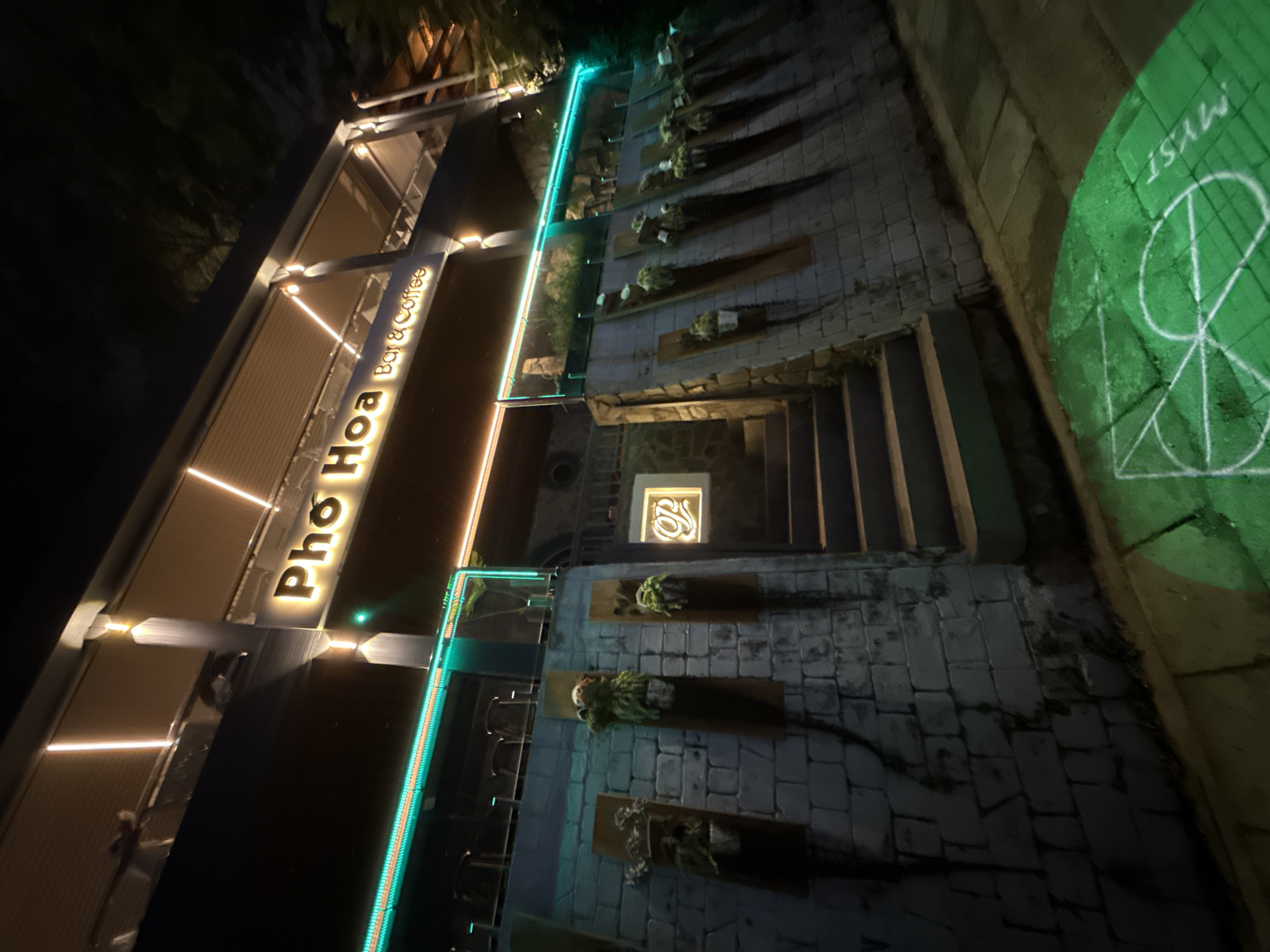 The image displays a nighttime view of an outdoor area, likely part of a restaurant or cafe in Da Lat, Vietnam. The scene is illuminated by artificial lights, creating a vibrant and welcoming atmosphere.
The prominent subject is a building with a large, glowing sign that reads "Pho Hoa Bar & Coffee" in yellow, sans-serif letters. The building's facade is decorated with horizontal warm white LED strip lights and a long, bright turquoise neon-style light strip running along its edge. Below the main sign, a smaller, square-shaped sign features a stylized white logo resembling an "S" or "JS" against a dark background.
A stone-paved pathway or steps are visible, leading downwards or across the frame. Along this path, rectangular wooden planters containing small green plants are mounted on a textured wall. On the far right, a bright green light is projected onto the stone ground, displaying the text "MYST" and a distinct geometric symbol that incorporates a circle and various straight lines, possibly a stylized letter or logo.
The time of day is clearly night, with the surrounding environment cloaked in darkness, making the artificial lighting the primary source of illumination. No people are present, allowing the architectural details and lighting design of the establishment to be the main focus. The overall impression is one of a modern and stylish bar or coffee shop designed for evening leisure.