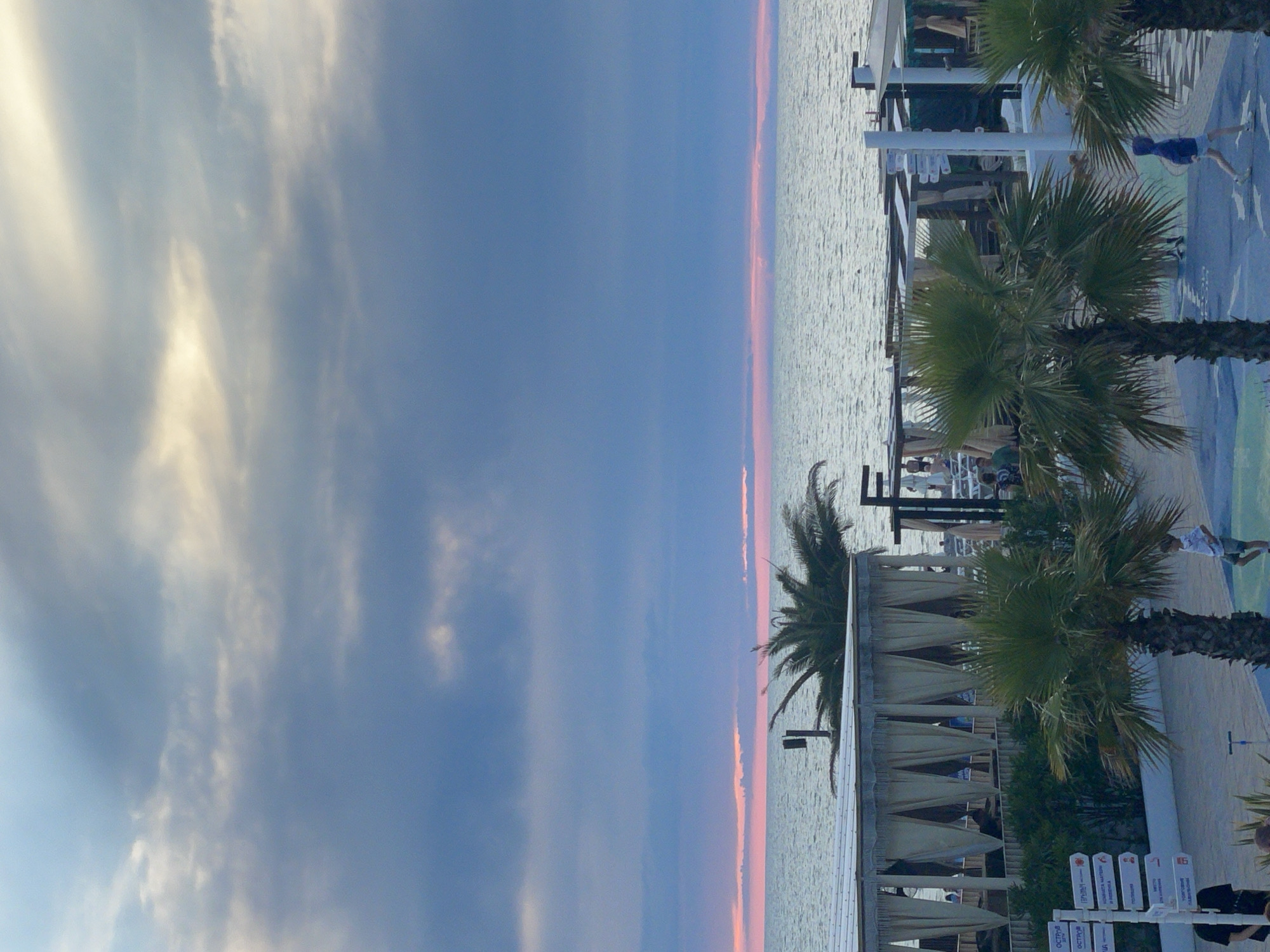 The image depicts a coastal scene, likely in Sochi, Russia, during either sunrise or sunset. The sky is filled with dramatic, layered clouds in shades of gray, blue, and hints of pink and yellow near the horizon. The sea is calm, reflecting the muted colors of the sky.
In the foreground, several palm trees are visible. To the left, a large open-air structure with white canvas coverings and curtains offers shade. It appears to be a dining or lounge area with people seated inside and near it. There are also directional signs with Russian text mounted on poles.
Further to the right, more open-air structures with white roofs and supports are set up. Several people are scattered throughout the scene. A young boy in a light-colored t-shirt and shorts is walking on a blue paved area. Further right, a person wearing a blue hoodie and shorts is walking. Some other individuals are visible in the background, some on lounge chairs near the beach.
The overall impression is a peaceful, seaside resort atmosphere, possibly a beach club or hotel area. The lighting suggests a transitional time of day, casting soft shadows and creating a serene ambiance.