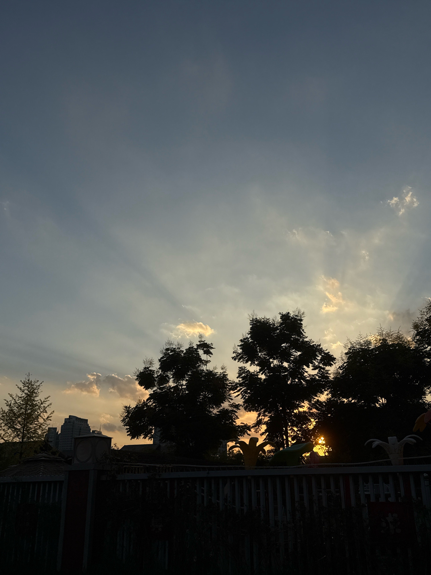 The image captures a sunset scene viewed from a fence, likely in Yiwu, China, given the presence of characteristic playground structures. The sky is a gradient of blue and grey, with the sun's rays breaking through scattered clouds, creating a dramatic crepuscular effect. Silhouetted against the sky are the dense foliage of several trees. In the mid-ground, beyond the trees, the outline of modern buildings is visible, suggesting an urban setting. The foreground is dominated by a white picket fence, with dark, shadowy vegetation growing behind it. Partially visible above the fence are abstract, colorful figures, possibly decorative elements from a playground or park. The overall impression is one of a tranquil evening in a city park.