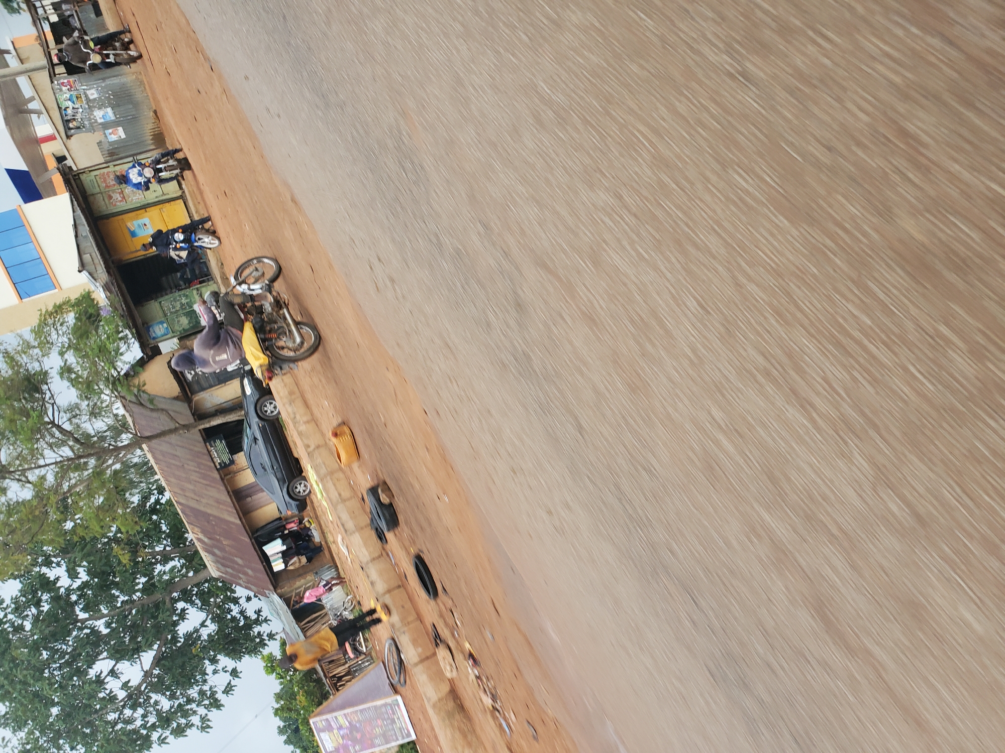 The image captures a bustling street scene in what appears to be a semi-urban or rural setting, oriented on its side. A wide, light brown, unpaved road dominates the right side of the frame, showing signs of dirt and light wear. Along the left side of the road are several simple, single-story structures, likely shops or homes. These buildings feature corrugated metal roofs and walls, with one structure displaying a distinct blue and white facade. Lush green trees flank the buildings on the far left, extending upwards towards an overcast or hazy sky.
Multiple subjects and objects populate the scene. Several motorcycles are visible; a prominent yellow motorcycle stands in the mid-ground, while another blue motorcycle is parked near one of the buildings. A black car is partially parked under a corrugated metal overhang. Several people are present, though their features are indistinct. One individual in dark grey and yellow attire is seen near the yellow motorcycle, and others are scattered near the buildings and along the roadside. Various items are strewn along the dirt shoulder, including what appears to be a black tire, a yellow jerry can, and other debris or goods. Posters are visible on some building walls, and a wooden stand with an illegible colorful sign is in the bottom left corner.
The scene depicts everyday activity, capturing a snapshot of daily life and commerce along the roadside. The time of day appears to be daytime, under an overcast sky. The overall atmosphere suggests a typical, calm day in the region, consistent with the provided location context of Vom, Nigeria.