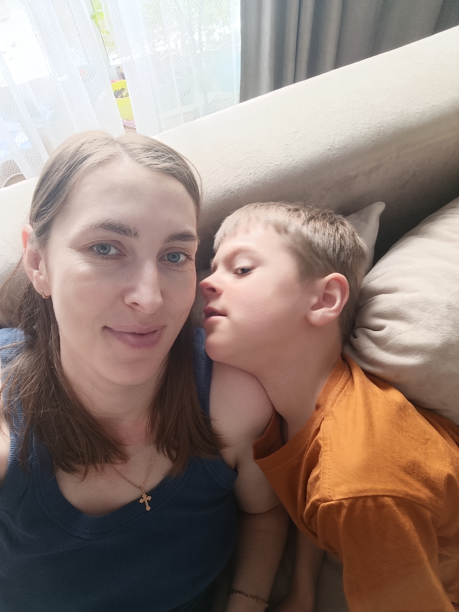 This close-up, selfie-style photograph captures an intimate moment between an adult woman and a young boy, likely a mother and child, in an indoor setting in Chaikovsky, Russia. The woman, with fair skin, light brown hair parted down the middle, and blue-grey eyes, is in the foreground, looking directly at the camera with a gentle, calm smile. She wears a dark blue tank top and a small gold cross pendant necklace. A small gold hoop earring is visible in her left ear.
To her right, the young boy, with light brown or blonde short hair, is leaning closely against her neck and shoulder. He wears an orange t-shirt and his face is turned slightly upwards and away from the camera, giving the impression of either resting or whispering. His expression is serene.
They are seated or reclining on a light-colored sofa or bed, part of a cream-colored pillow visible behind the boy. The background reveals a bright window covered with sheer white curtains, with heavier grey curtains to the right. Hints of greenery are visible outside the window, suggesting a daytime setting with natural light illuminating the scene. The overall mood is one of warmth, affection, and relaxation. No visible text is present in the image.