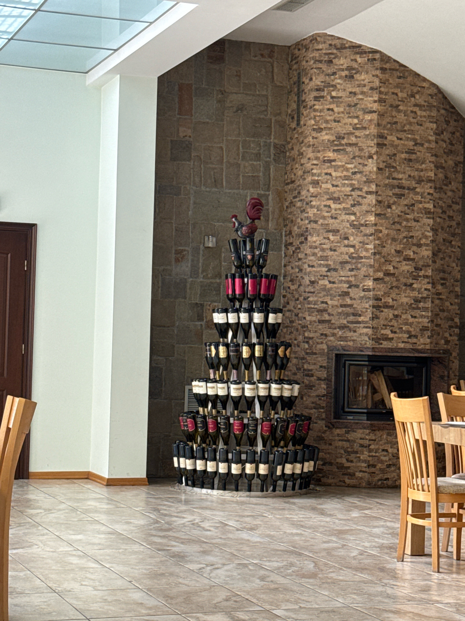 The image depicts an indoor scene, likely a restaurant or wine cellar, with a distinctive wine bottle display. The display is arranged in a tiered, conical shape, resembling a Christmas tree, and is adorned with numerous wine bottles of various colors, predominantly dark glass with some red-necked bottles. At the very top of the display, a small, decorative rooster figure is perched.
The background features a large fireplace with a dark metal insert and visible logs. The fireplace is surrounded by a wall clad in textured, rustic-looking stone or brick tiles. To the left of the wine display, a light-colored wall and a dark wooden door are visible. The ceiling appears to be a combination of a white surface and a section of glass panels, possibly a skylight. The floor is tiled with large, light-colored square tiles.
In the foreground and to the right, part of a dining area is visible. Two wooden chairs with light upholstery are positioned around a wooden table.
The scene is static, with no people actively engaged in any activity. The lighting suggests ambient indoor light, with some natural light possibly filtering through the glass ceiling panels. The overall impression is one of a decorative display within a hospitality establishment. While the location is stated to be Ustina, Bulgaria, there are no specific visual cues within the image that definitively identify this geographic location.