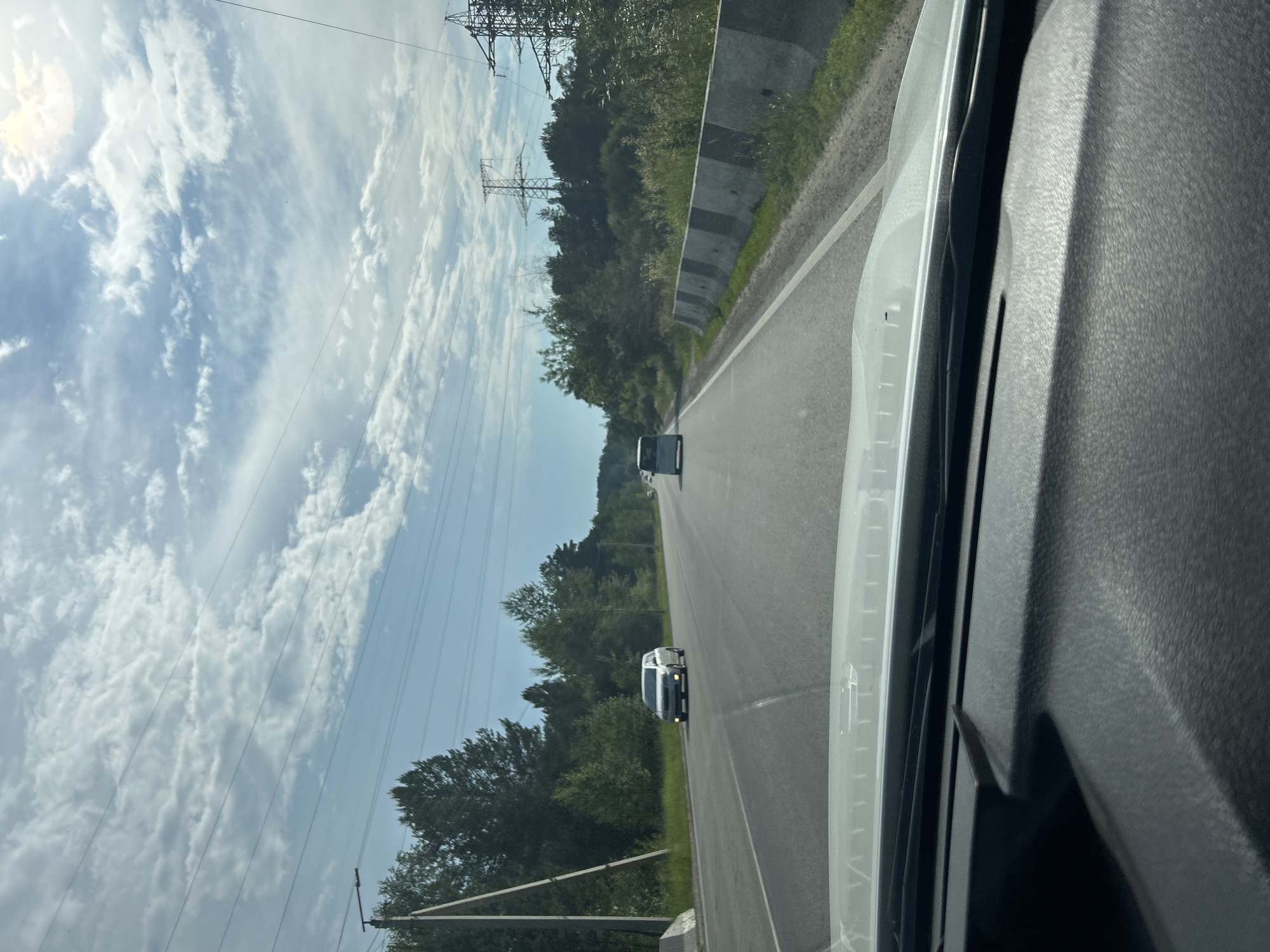 The image depicts a view from inside a vehicle traveling on a highway near Angarsk, Russia. The road is asphalt, with two lanes visible in the direction of travel and a concrete barrier separating the lanes from the opposite direction. Trees and vegetation line the sides of the road, and tall power lines and transmission towers are visible in the distance. The sky is partly cloudy, with the sun appearing to be high in the sky. Two vehicles are visible on the road ahead, one silver sedan in the left lane and a white van in the right lane. The interior of the vehicle, including the dashboard and windshield, is partially visible in the foreground.