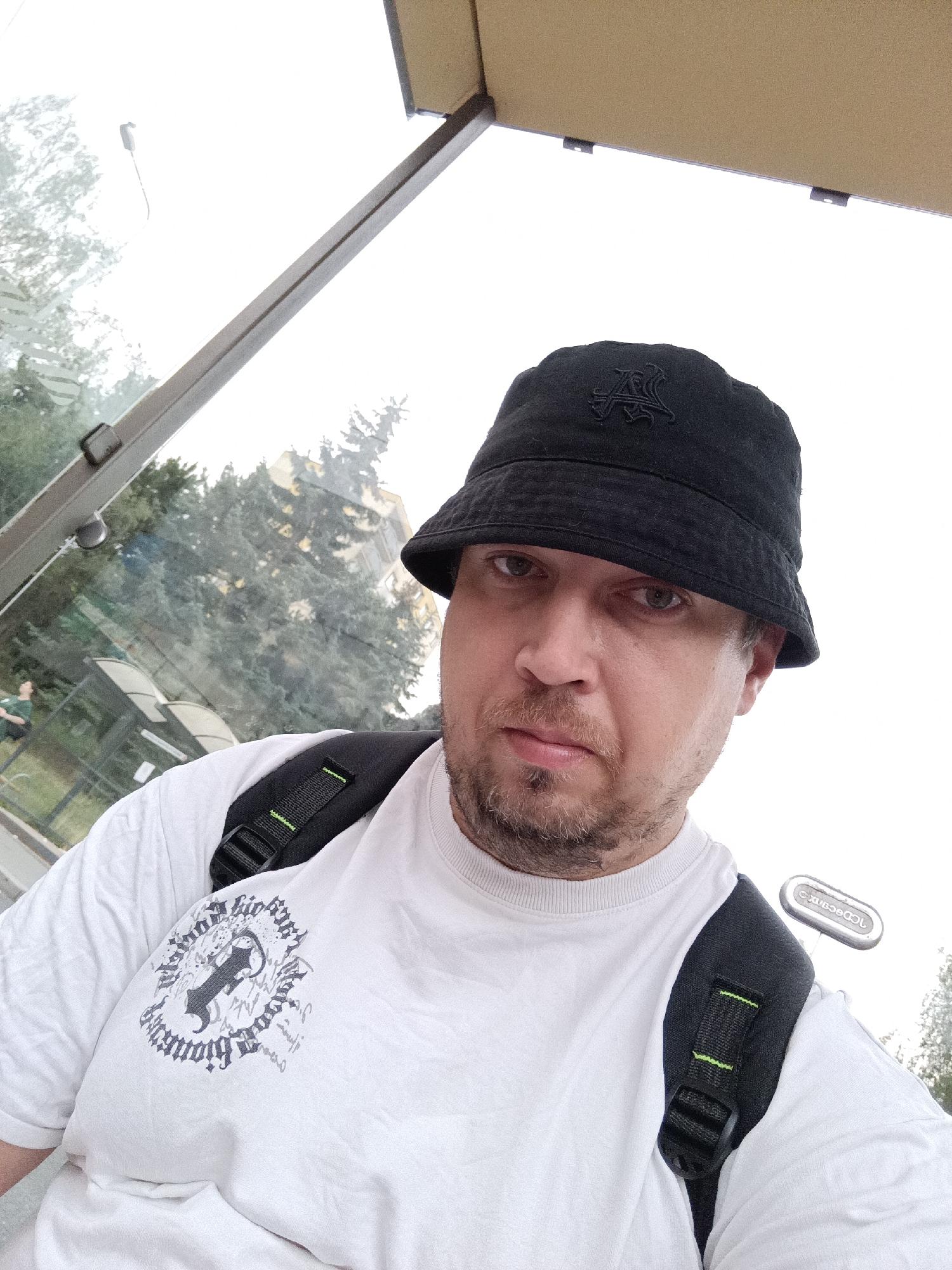 The image is a close-up, selfie-style shot of a man wearing a black bucket hat with an embroidered logo and a white t-shirt with a graphic design. He is also wearing a black backpack with green accents. The man has a beard and is looking directly at the camera. The background appears to be a bus stop with a glass enclosure, through which trees and a building are visible. The lighting suggests an overcast day. The location is identified as a bus stop in Prague, Czechia.