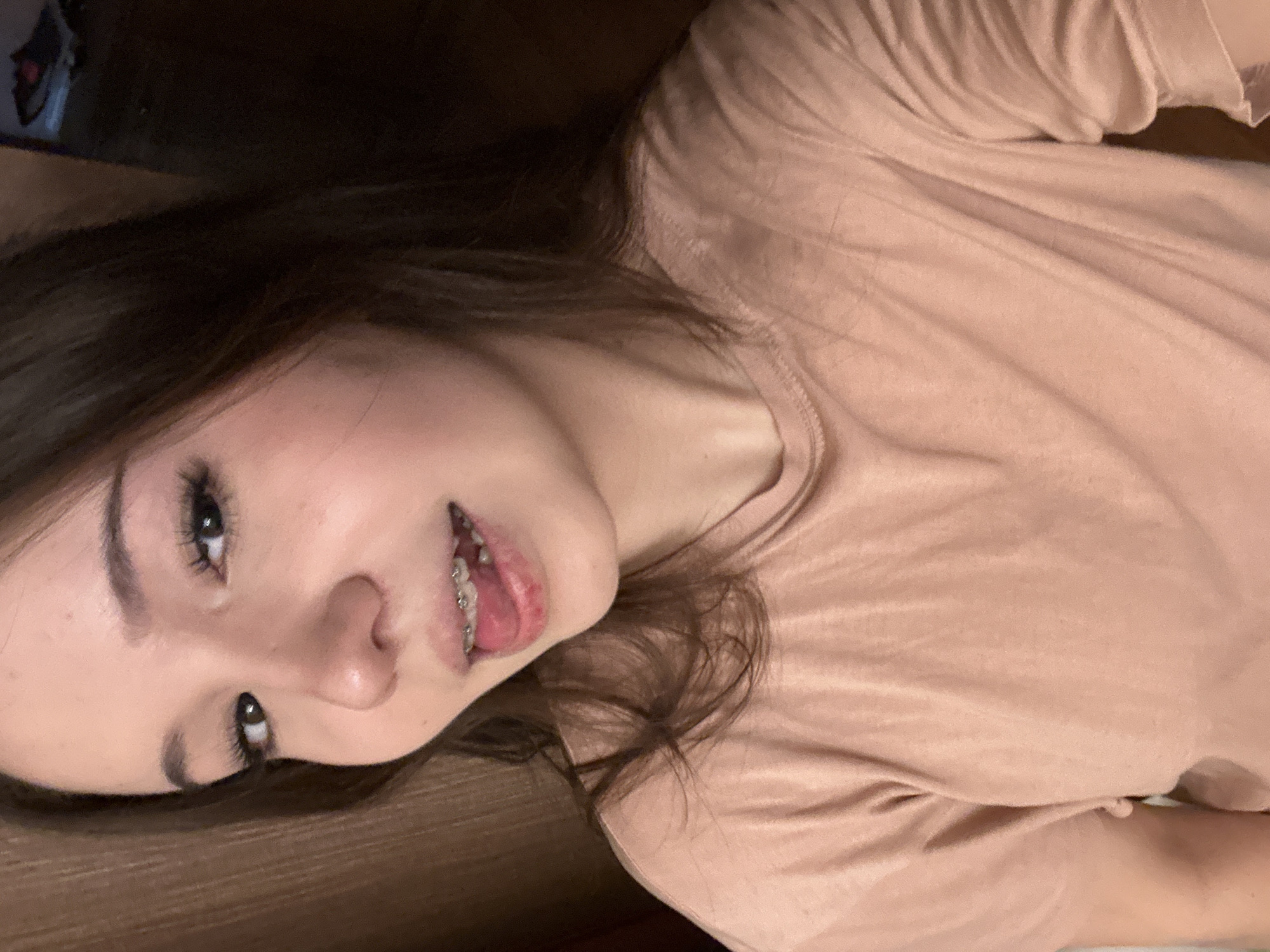 The image is a close-up, rotated portrait of a young woman taken indoors, likely a selfie. She has dark brown hair that frames her face and flows down her shoulders. Her skin tone is fair, and she has well-defined dark eyebrows and long, dark eyelashes highlighting her light-colored eyes. She is wearing a plain, light pink or peach-colored short-sleeved t-shirt, which shows some soft wrinkles in the fabric.
The woman's head is tilted, and she is looking directly into the camera with a playful expression. Her mouth is slightly open, revealing braces on her upper teeth, and her tongue is partially visible, touching her upper lip or braces.
The background is dark and appears to be a solid, textured surface, possibly wood or a dark wall. There are no distinct objects or features in the background other than this dark surface, which provides a simple contrast to her lighter skin and shirt. The lighting is soft and appears to be artificial, creating subtle highlights on her face and casting gentle shadows. No visible text is present in the image. The overall impression is casual and intimate.