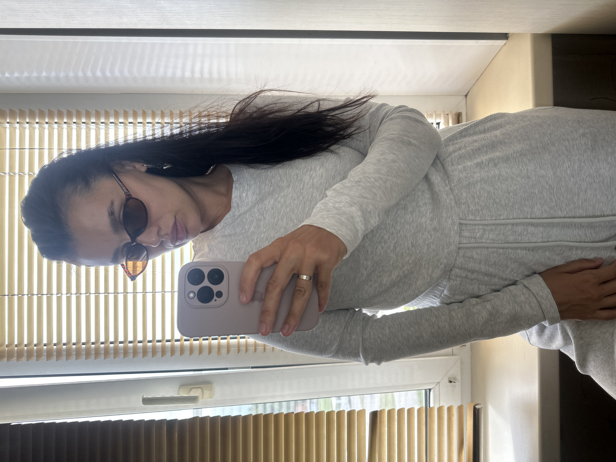 A woman is taking a selfie in a room. She is wearing a light gray loungewear set and sunglasses. She is holding a phone in her right hand, and a ring is visible on her left ring finger. The background consists of blinds on a window and a wall. The image does not provide any specific information about the city of Chaikovsky, Russia.