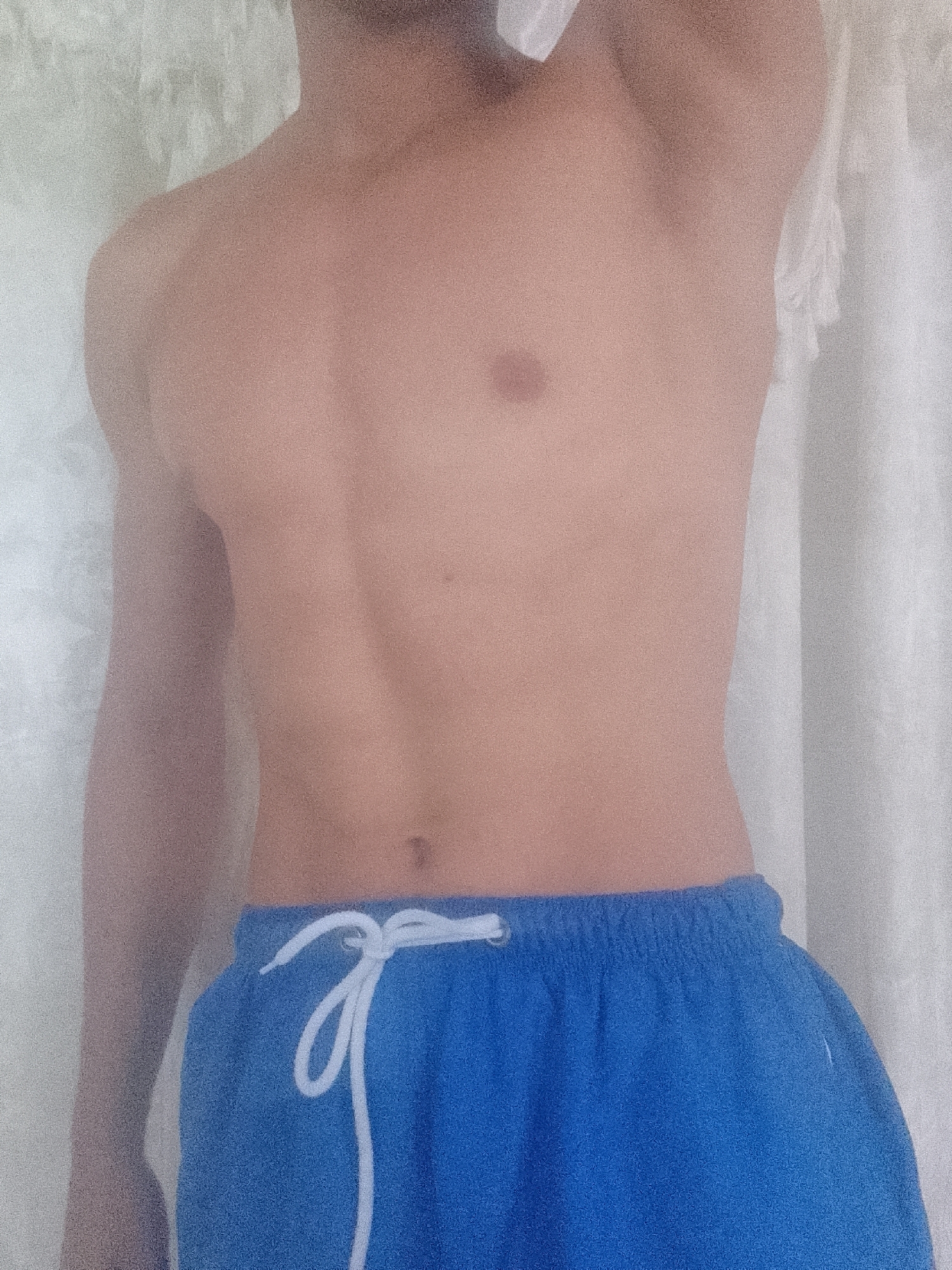 A close-up shot depicts the torso and upper thighs of a light-skinned male figure against a blurry white background. He is shirtless and wearing blue swim trunks with a white drawstring. No other identifying features are visible. The location in Juan Griego, Venezuela, is not discernible from the image.