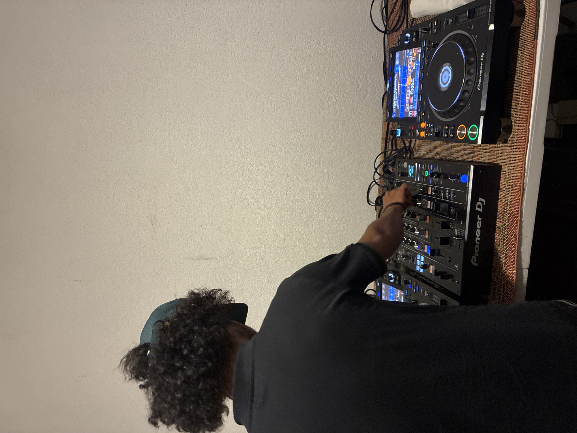 A person with curly hair, wearing a dark shirt and a baseball cap, is operating a Pioneer DJ mixer and CDJ player setup. The equipment is placed on a table with a patterned tablecloth, set against a light-colored wall. The exact location in Mexico City, Mexico, cannot be determined from the image alone.
