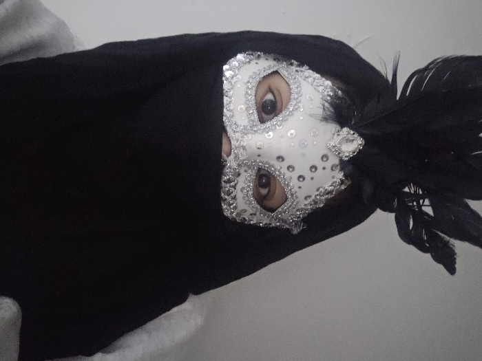 A person is wearing a white Venetian mask adorned with silver sequins and black feathers. The mask covers the upper portion of their face, revealing only the eyes. The person is draped in a black garment with a hint of a white or grey fabric underneath. The background appears to be a plain white or light-colored wall. The image is slightly tilted to the right.