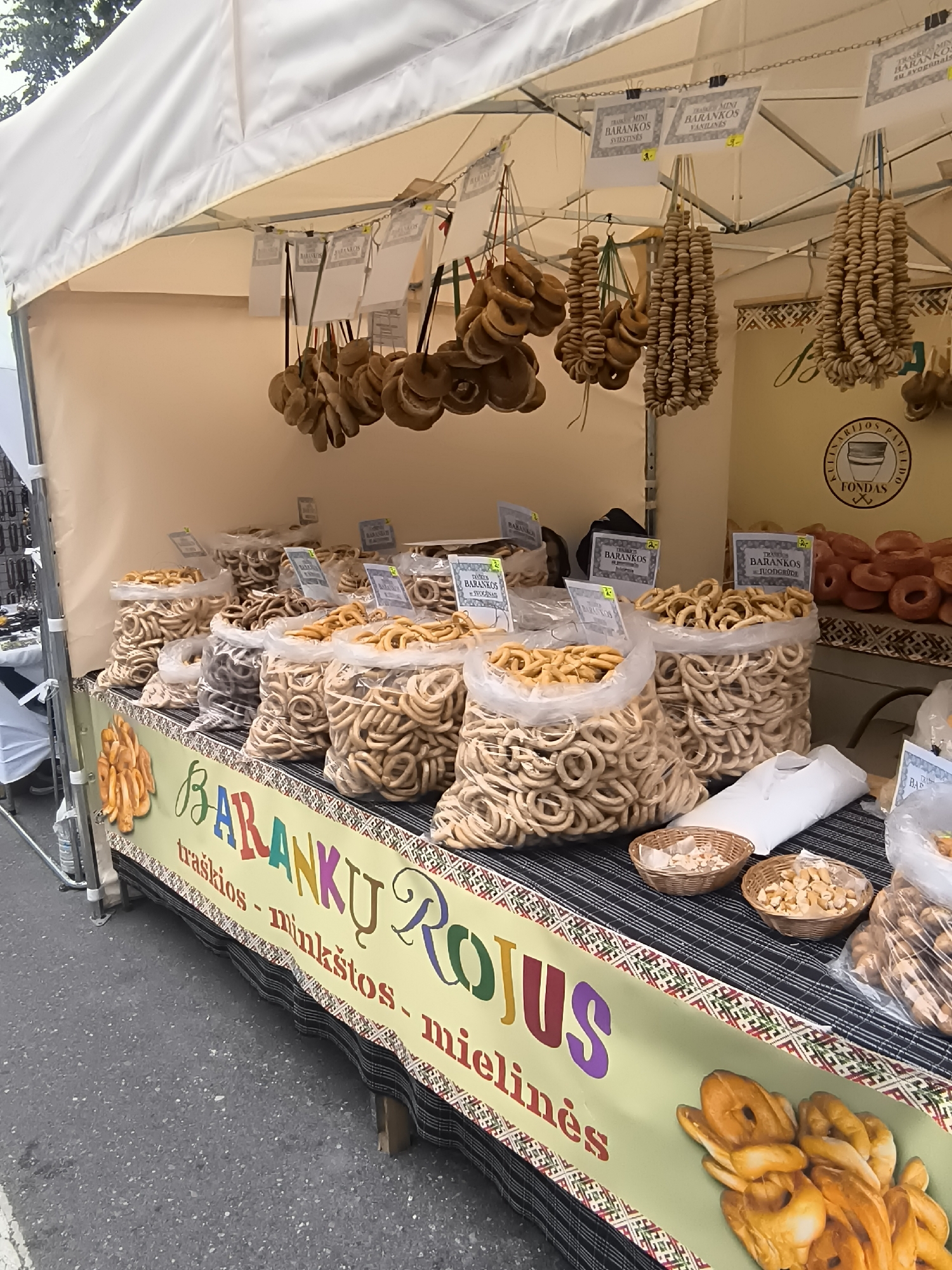 The image depicts a food stall at an outdoor market in Klaipėda, Lithuania, specializing in ring-shaped baked goods, likely traditional Lithuanian 'barankos'. The stall is covered by a white tent. In the foreground, a table is laden with large plastic bags filled with these ring-shaped pastries. Some are light brown, while others appear darker. Woven baskets containing smaller pieces of baked goods are placed on the table. A colorful banner runs along the front of the table, featuring the text "BARANKŲ ROJUS" in large, multicolored letters, followed by "traškios - minkštos - mielinės" which translates to "crunchy - soft - honeyed". The banner also has a graphic of several larger, braided pastries. Behind the table, more bags of baked goods are visible, and some are hanging from the tent structure, displayed in bunches. Small signs with Lithuanian text, likely prices or descriptions, are attached to the hanging pastries. A circular logo with a stylized illustration of a bread oven and the text "KULINARIJOS FONDAS" is visible on the right side of the stall. The ground surface in front of the stall appears to be asphalt, suggesting a street or public square setting. No people are clearly visible interacting with the stall. The overall impression is a vibrant display of traditional Lithuanian baked goods at a marketplace.