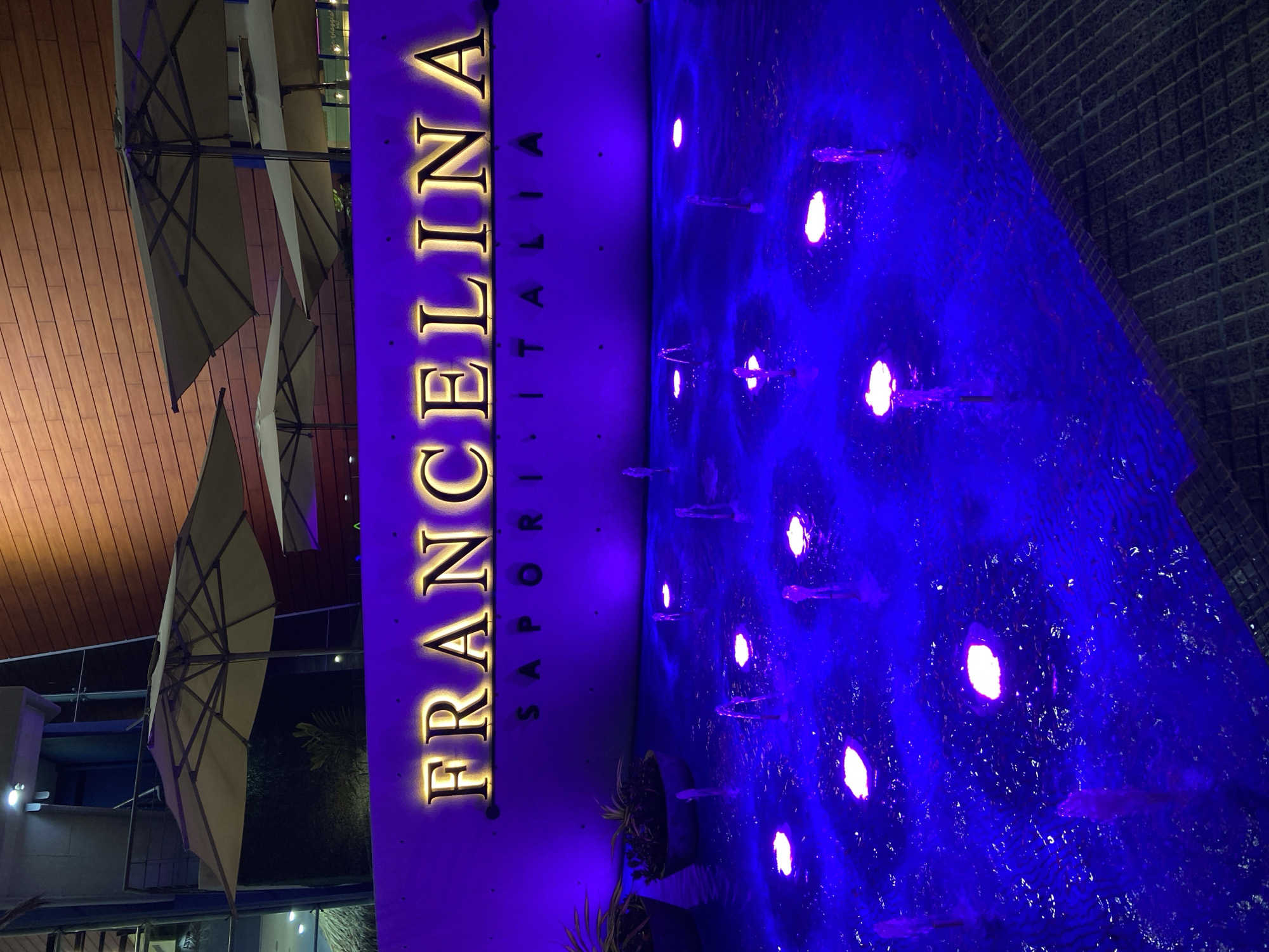 In Porlamar, Venezuela, a restaurant named FRANCELINA SAPORI D'ITALIA is illuminated by purple and yellow lights. The restaurant's name is displayed in large, illuminated gold letters on a dark wall above a water feature. The water feature has multiple jets of water and purple lights that illuminate the water from below, creating a vibrant, aquatic display. Surrounding the restaurant, there are large, neutral-colored umbrellas providing shade. The building behind the umbrellas has a textured, wooden facade and a dark upper section with visible lighting fixtures. In the foreground, planters with green foliage are visible. The overall scene suggests an outdoor dining or entrance area of a restaurant at night.