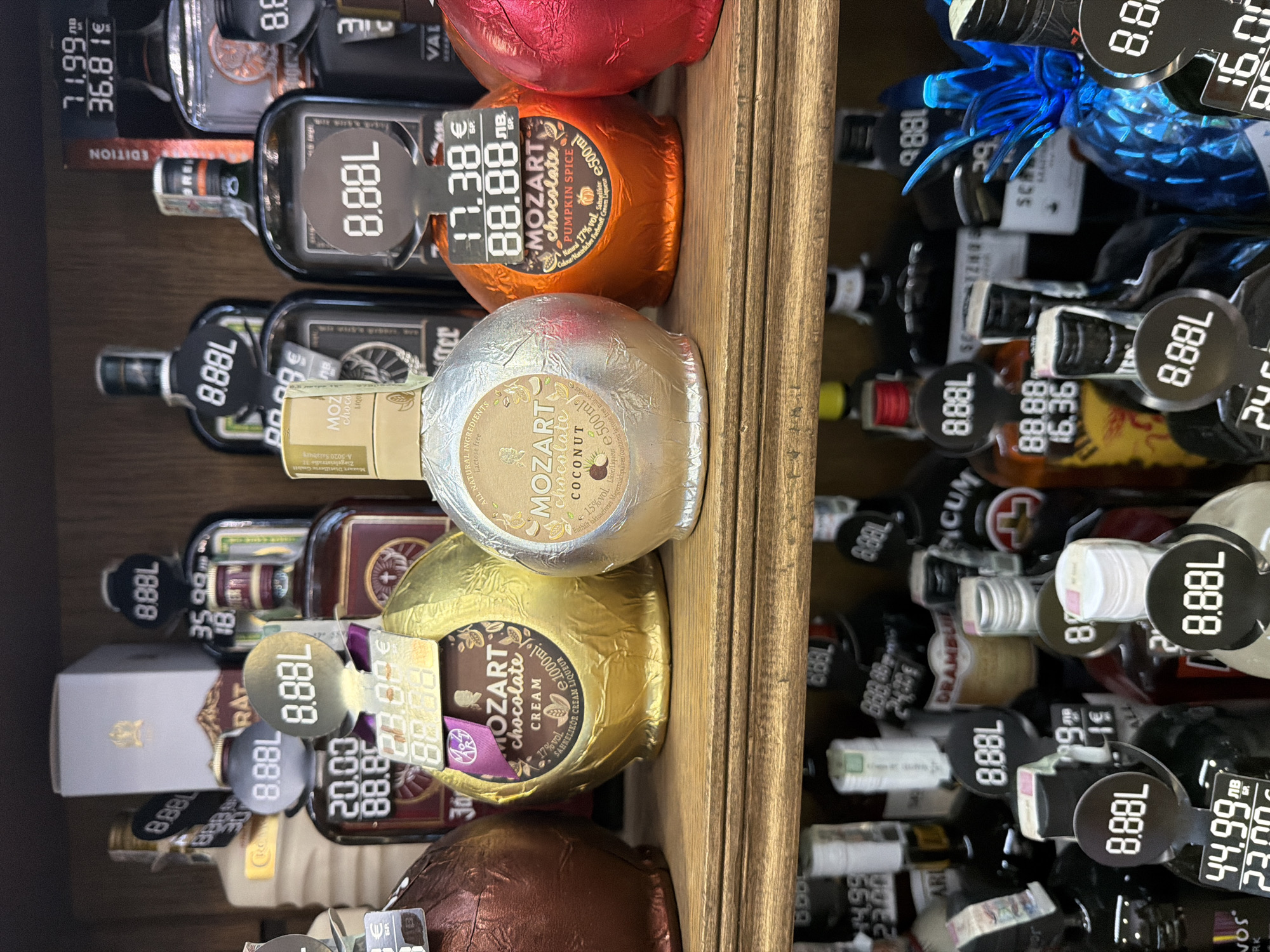 The image shows a shelf display of various bottles of alcohol, primarily liqueurs, in a retail environment. The bottles are arranged on two wooden shelves. The upper shelf displays several bottles, including what appear to be Jägermeister and Mozart Chocolate Liqueur in distinctive spherical packaging. Some bottles have price tags and volume indicators, such as "8.88L" and prices in Euros and Lev. The lower shelf shows a wider variety of bottles, many with "8.88L" tags, and some with prices like "44.99 лв" and "23.00 лв". The overall lighting is somewhat dim, suggesting an indoor setting. The style of the displays and the pricing in Lev strongly suggest this is a liquor store or a duty-free shop in Bulgaria. Given the currency and the likely context of such a display, this scene is likely located in a retail establishment in Sofia, Bulgaria. There are no people visible in the image.