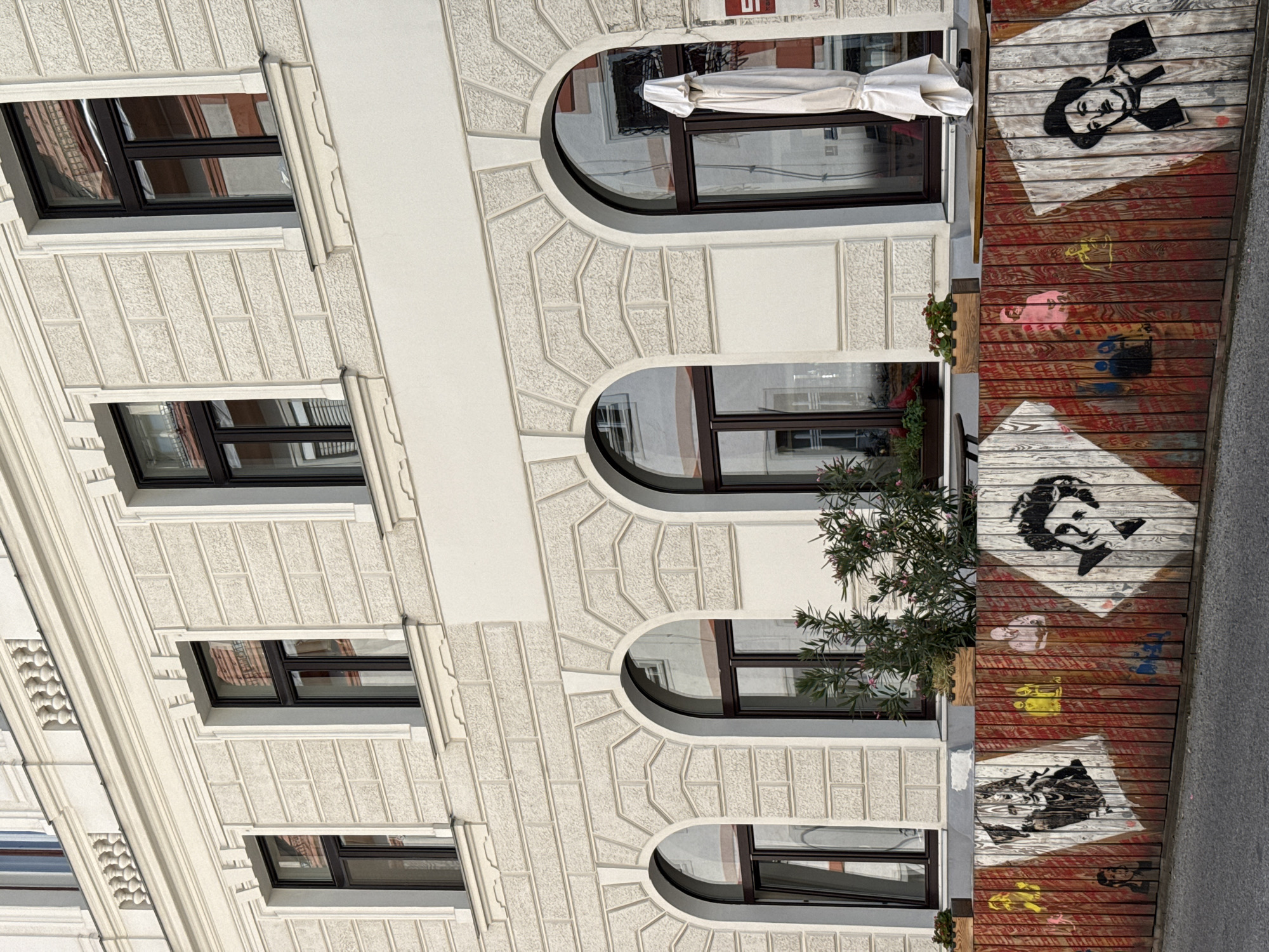 The image captures the facade of a building with classical architectural elements in Vienna, Austria, specifically in front of what appears to be a cafe or restaurant. The building features a pale, textured facade with decorative molding around arched windows. Several windows are visible, some reflecting the surrounding environment.
In the foreground, a wooden barrier serves as a decorative element for an outdoor seating area. This barrier is adorned with stenciled portraits, primarily of famous figures, rendered in black and white with some colored accents like yellow and pink. The style of the portraits suggests iconic personalities, possibly from film or history. One prominent portrait is of Charlie Chaplin, recognizable by his hat and mustache. Other figures are also depicted with distinct features.
Behind the barrier, tables and chairs are set up, indicating the space is an outdoor extension of a business. A white, closed umbrella stands near one of the tables, suggesting it's either not in use or the weather is not requiring it. Greenery, including small plants and flowers in wooden planters, adds a touch of nature to the scene. The overall impression is one of a charming urban setting with an artistic touch.