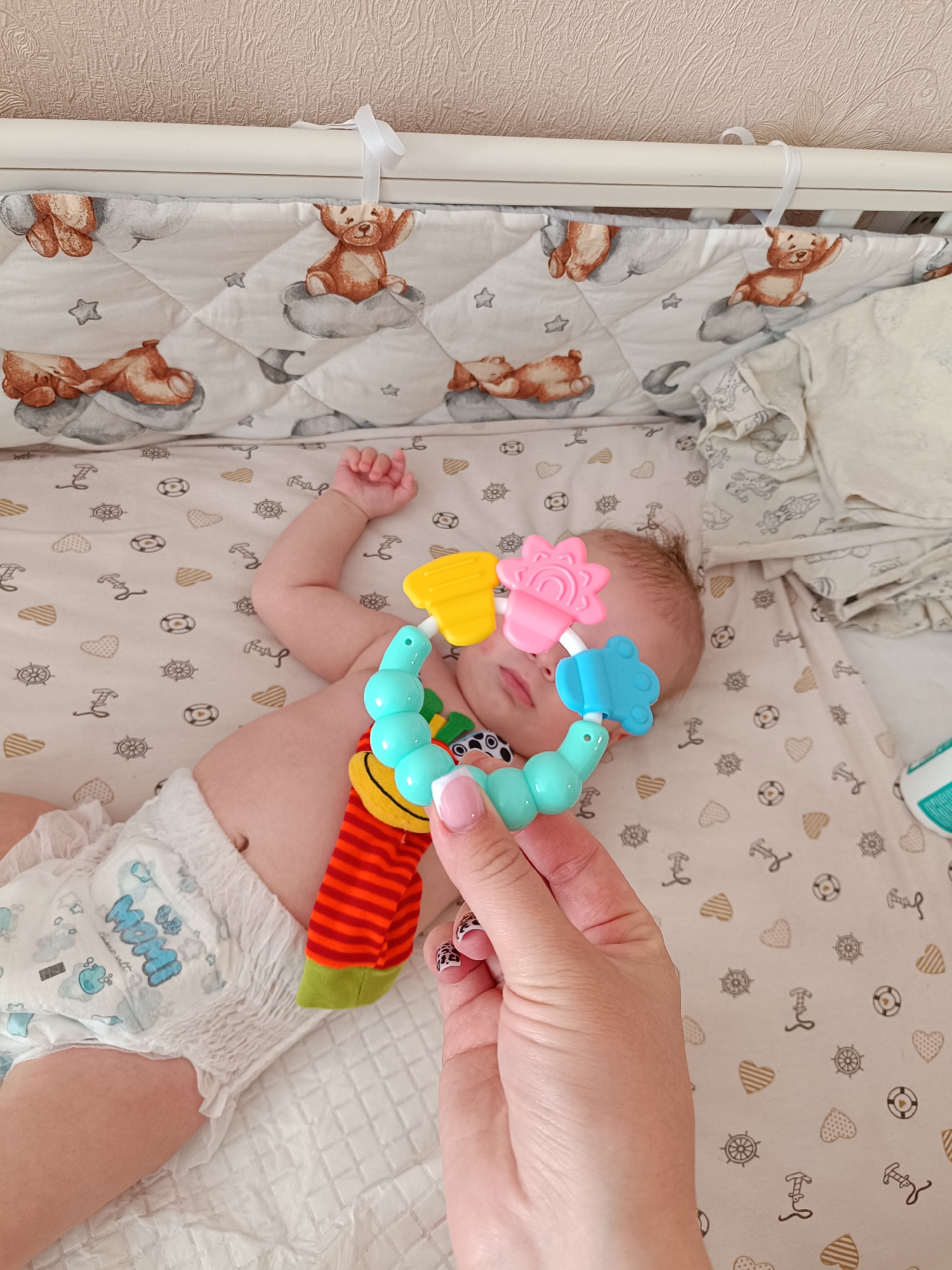 This vertical shot captures an intimate indoor scene, likely in a nursery or bedroom in Tobolsk, Russia. A young infant lies on their back in a crib, wearing a white disposable diaper with blue "MoM" text and cartoon frog-like characters. Their feet are covered by vibrant red and orange striped socks with a green tip. The baby's gaze is directed towards a colorful rattle/teether toy held by an adult's hand.
The adult's hand, visible in the lower right foreground, has manicured nails featuring a French tip and a leopard print design. The toy is brightly colored in turquoise, yellow, pink (shaped like a flower), and light blue (shaped like a car).
The crib is outfitted with a light-colored sheet patterned with maritime motifs such as ship wheels and anchors, interspersed with striped hearts. The crib's side is lined with a quilted bumper pad adorned with illustrations of brown teddy bears on clouds, alongside stars and crescent moons. A white ribbon is tied to the white crib rail. In the background, a light-beige textured wallpaper is visible. To the right, folded white blankets or fabric are stacked, and a small white and green bottle is partially visible at the edge of the frame. The scene portrays a tender interaction between the adult and the baby.