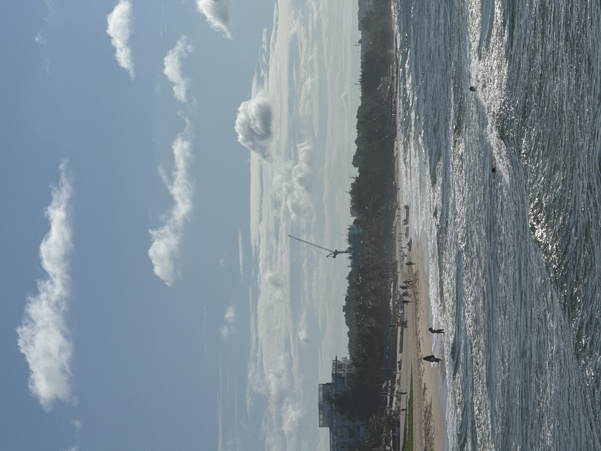 The image captures a coastal scene in Phan Thiết, Vietnam, with a view of the ocean, beach, and a town in the background. The foreground is dominated by the ocean, with choppy waves rolling towards the shore. Several people are visible in the water, some appearing to be swimming or wading. Further back, the sandy beach is populated with individuals walking and relaxing. Beach umbrellas and some watercraft are scattered along the shoreline. In the middle ground, a dense line of palm trees separates the beach from the town. Buildings, including a large multi-story structure and a construction crane in the distance, are visible behind the trees, suggesting ongoing development or infrastructure projects. The sky above is a clear blue with scattered white clouds, indicating a bright, possibly sunny day.