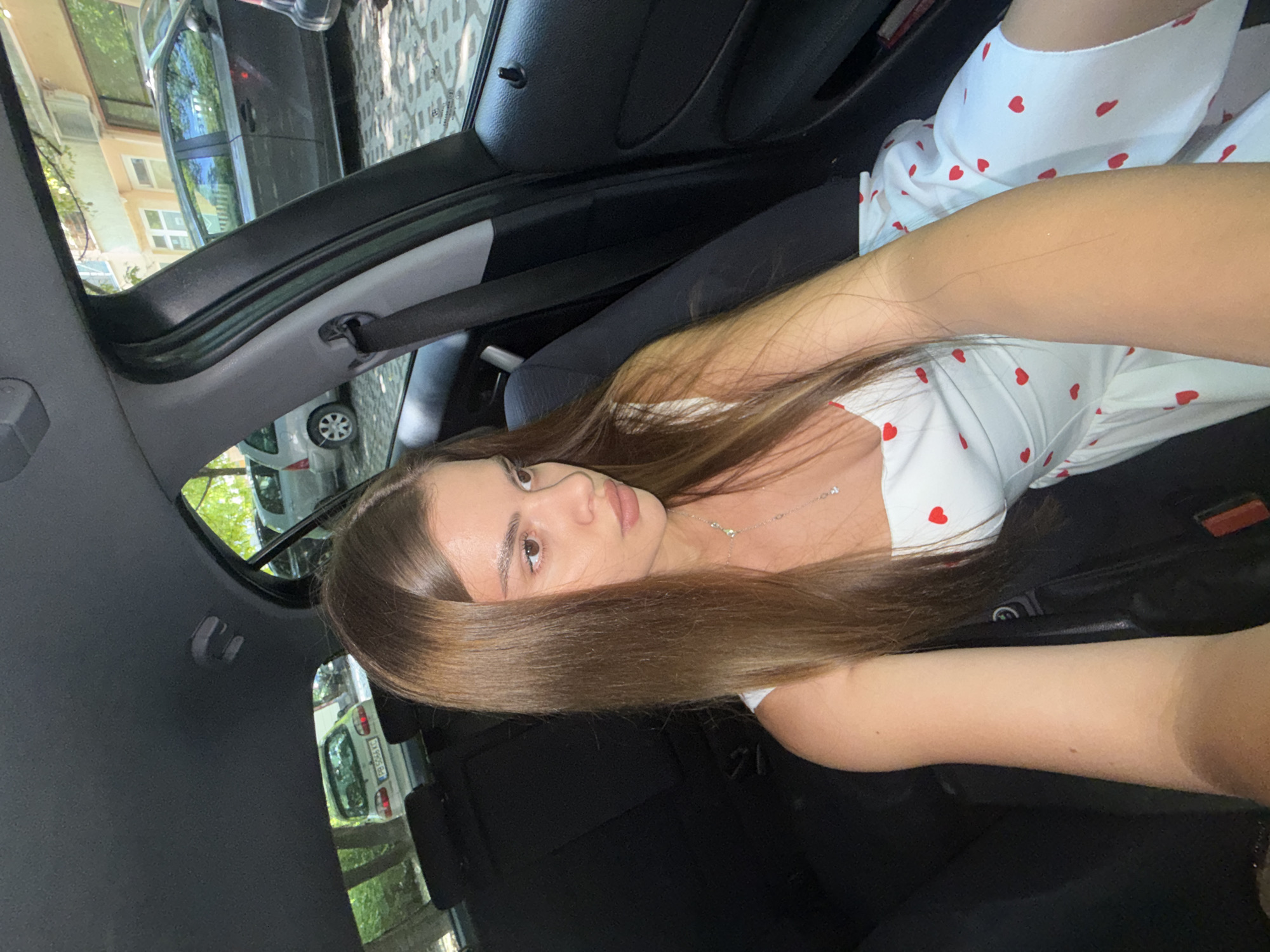 A young adult female is seated in the front passenger seat of a dark-interior vehicle, holding a camera or phone with her extended right arm to capture a selfie. She has long, straight, brown hair and is wearing a white dress adorned with small red heart patterns and a slender, silver-toned necklace. Her gaze is directed away from the camera, towards the right side of the frame. Through the rear window of the vehicle, a white hatchback is visible in the background, displaying a Bulgarian license plate "PB 5066 XC," confirming the location as Plovdiv, Bulgaria. The exterior environment includes a cobblestone or patterned paved street, various parked automobiles including a dark grey SUV/crossover, buildings with windows and an external air conditioning unit, and trees with green foliage. The scene is illuminated by natural daylight.