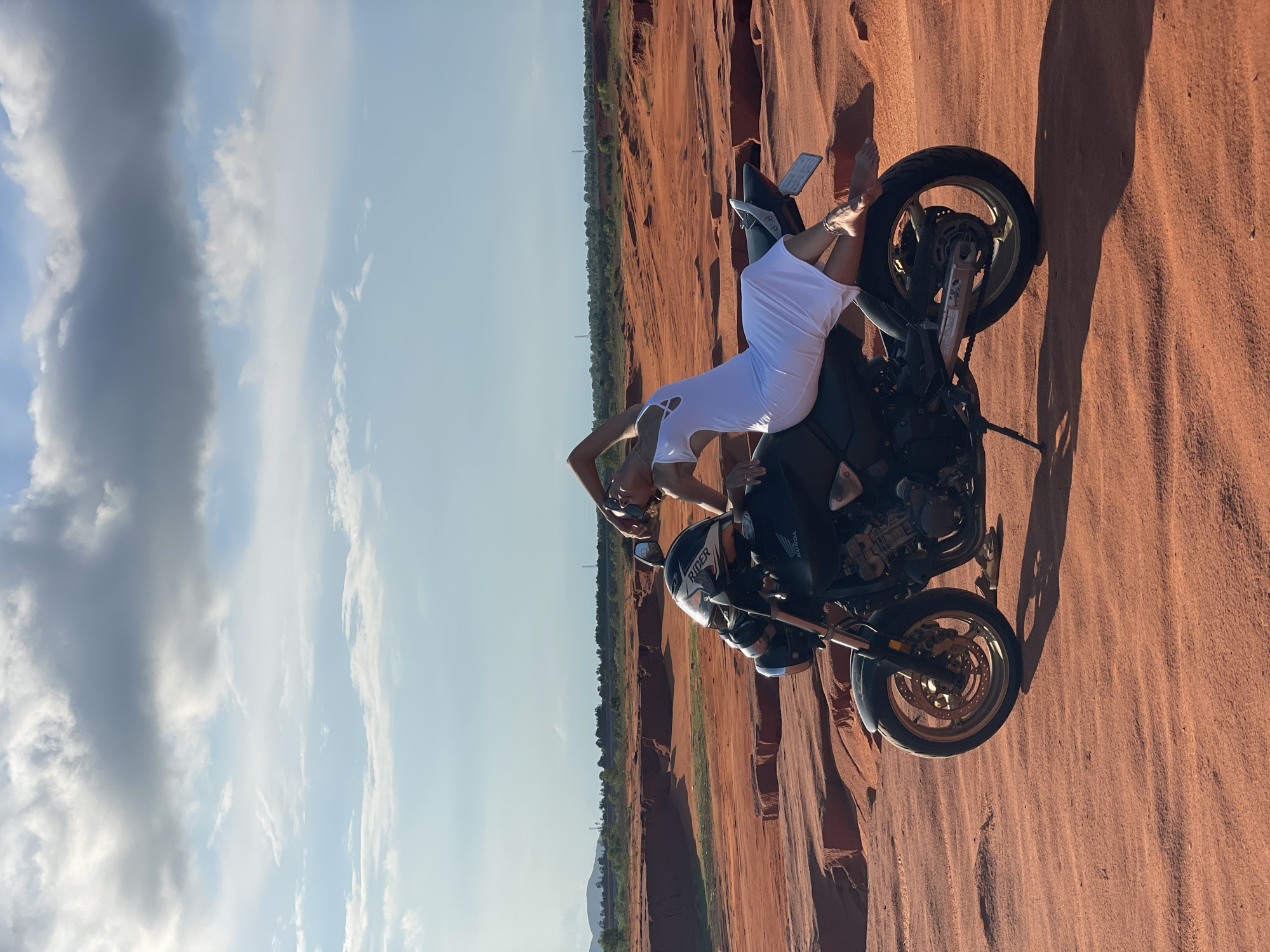 In Mui Ne, Vietnam, a person in a white dress reclines on a black Honda motorcycle parked on a sandy, red-orange terrain. The motorcycle faces left, with its front wheel slightly to the left of the image center. The person is positioned on the seat, leaning back with their arms behind their head, legs extended towards the rear of the motorcycle. Their body is arched, creating a dynamic pose against the arid landscape. The background features sculpted, reddish-brown rock formations and a distant line of trees and hills under a partly cloudy sky. The sky is a gradient of pale blue to white, with fluffy white and darker gray clouds.