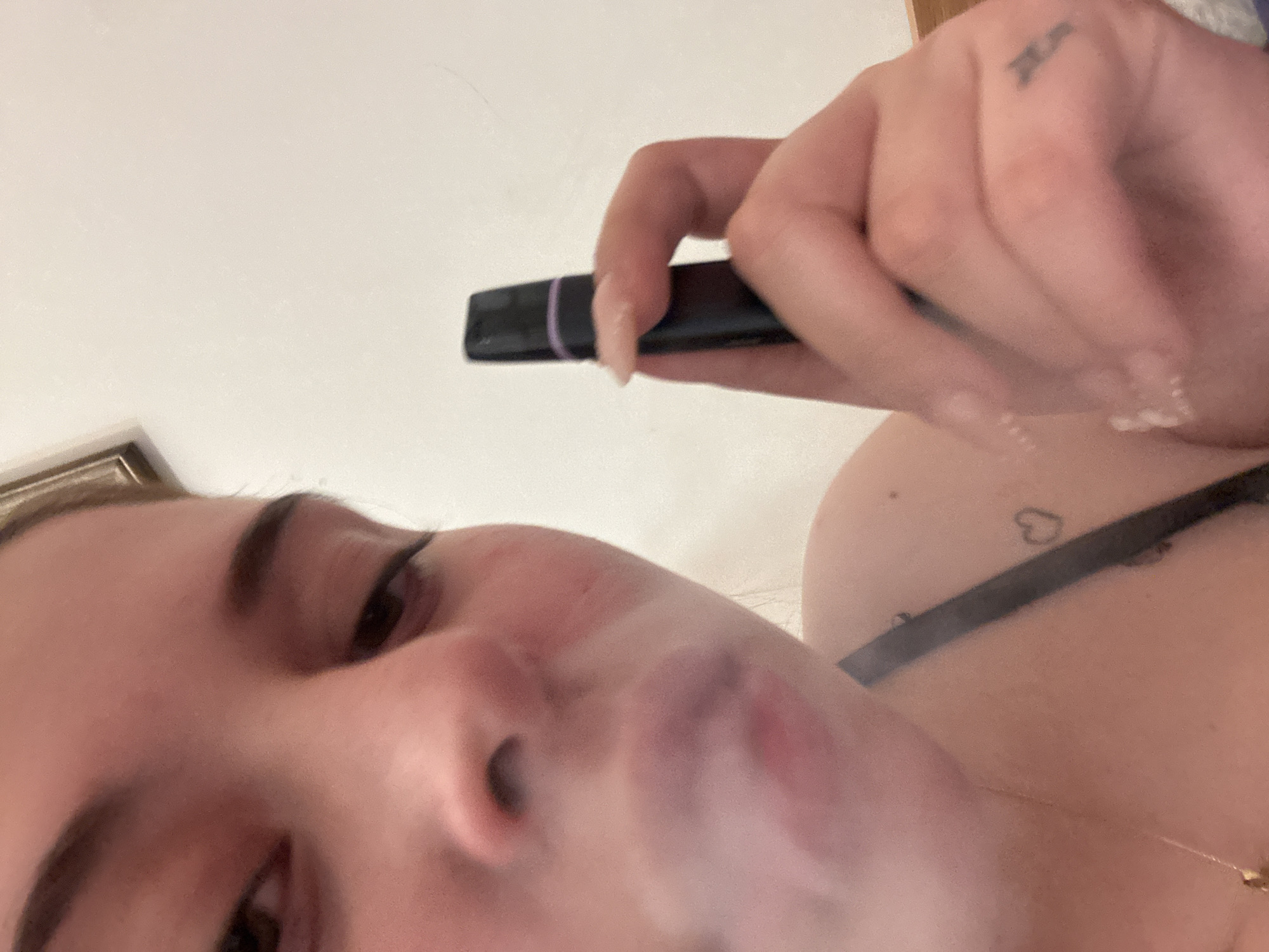 A close-up, tilted, low-angle view captures a person exhaling vapor from a vaping device. The person is holding a black vaping device with a purple band. The person's left eye and eyebrow, nose, and part of their cheek are visible. Vapor is emanating from their mouth. A hand holding the device is in the upper right of the frame with short fingernails. On the hand is a tattoo. In the upper right shoulder area, there is a small heart tattoo visible along with a bra strap and a gold necklace. The background is a plain, light color. The image does not contain any information to suggest the location is Troyan, Bulgaria.