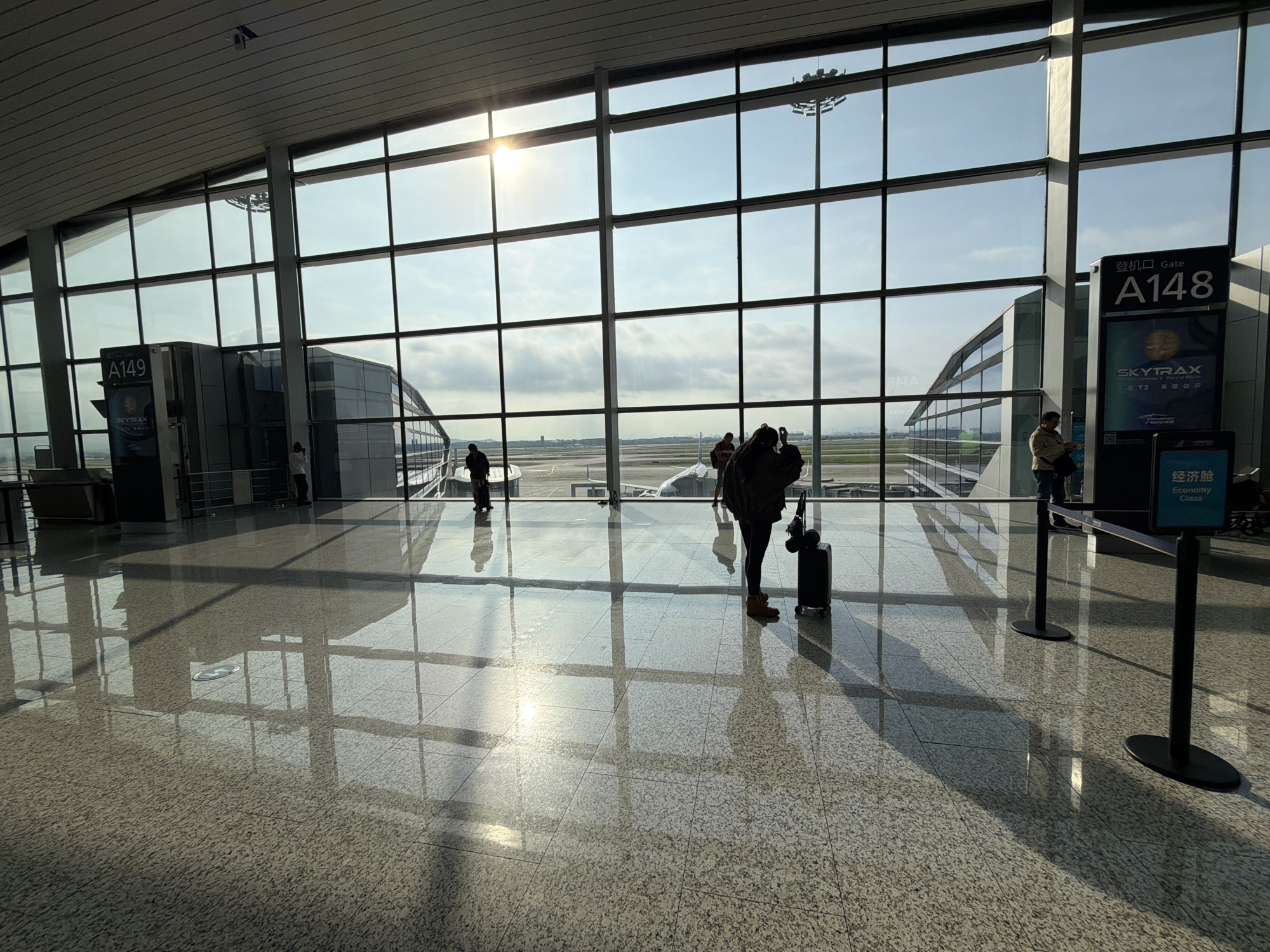The image depicts the interior of an airport terminal. Large windows offer a view of the airfield, where airplanes are parked. Several individuals are present in the terminal. A person is standing near a suitcase. Another person is taking pictures. There is a sign indicating gate A148. The location is likely Guangzhou Baiyun International Airport, in the Baiyun District, China. The floor is shiny, reflecting the light and shapes from the windows and terminal structure.