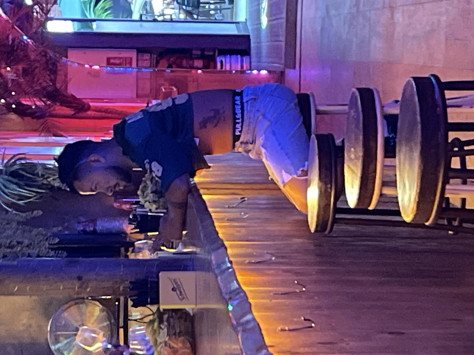 A man with dark hair and a beard sits at a wooden bar. He is wearing a blue and green cropped t-shirt, light blue ripped jeans, and a black belt with the text "PULL&BEAR" on it. He has a tattoo on his stomach. The bar is decorated with orange lights along its edge. There are several stools at the bar, and the man is sitting on one of them. In the background, there is a fan, bottles, and plants. The scene appears to be in a bar or restaurant.