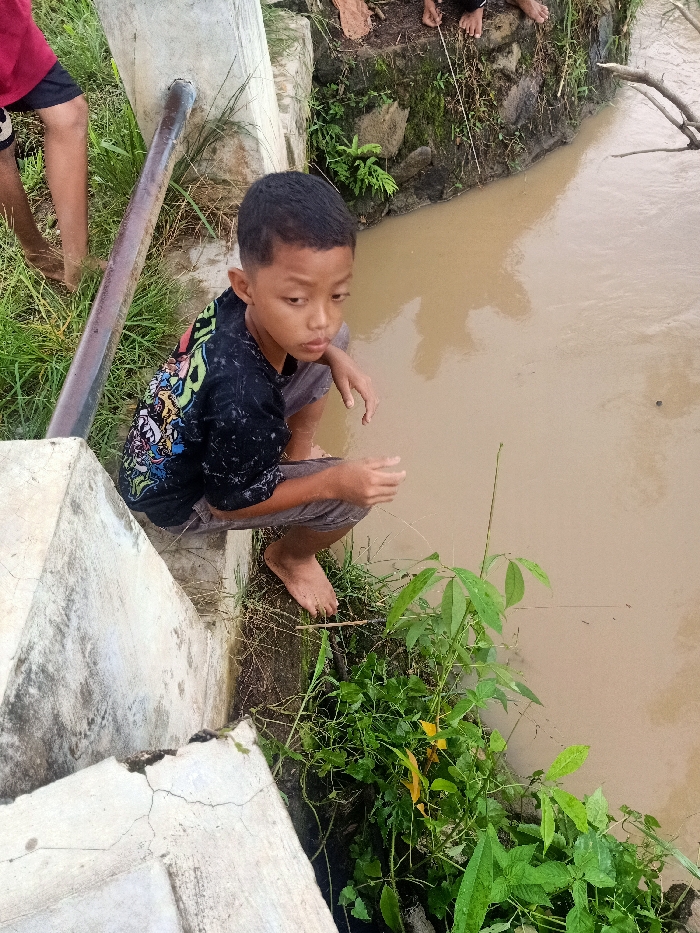 A young boy with short dark hair, wearing a black t-shirt with colorful graphics and grey shorts, is seen crouching barefoot on a concrete structure next to a muddy brown body of water, likely a river or canal, in Pubian, Indonesia. His gaze is fixed intently on the water below, and his hands are positioned as if he is holding something or preparing to engage with the water, possibly fishing or observing aquatic life.

To the left of the boy, the bare legs and feet of another person are visible standing on grassy ground near a metal pipe that runs alongside the concrete structure. Further in the background, near the opposite bank of the waterway, the feet of additional individuals can be seen, suggesting others are present in the vicinity.

The water appears murky and brown, reflecting the overcast sky or indicating a muddy bottom. A large tree branch is partially submerged in the water on the far side. Lush green vegetation, including grass and various leafy plants, grows along the edges of the concrete structure and the riverbank. The concrete structure itself shows signs of wear with some cracks. The scene depicts a casual outdoor activity by the water, possibly fishing or simply observing nature. The overall atmosphere appears calm and focused on the interaction with the natural environment. No visible text is present in the image.