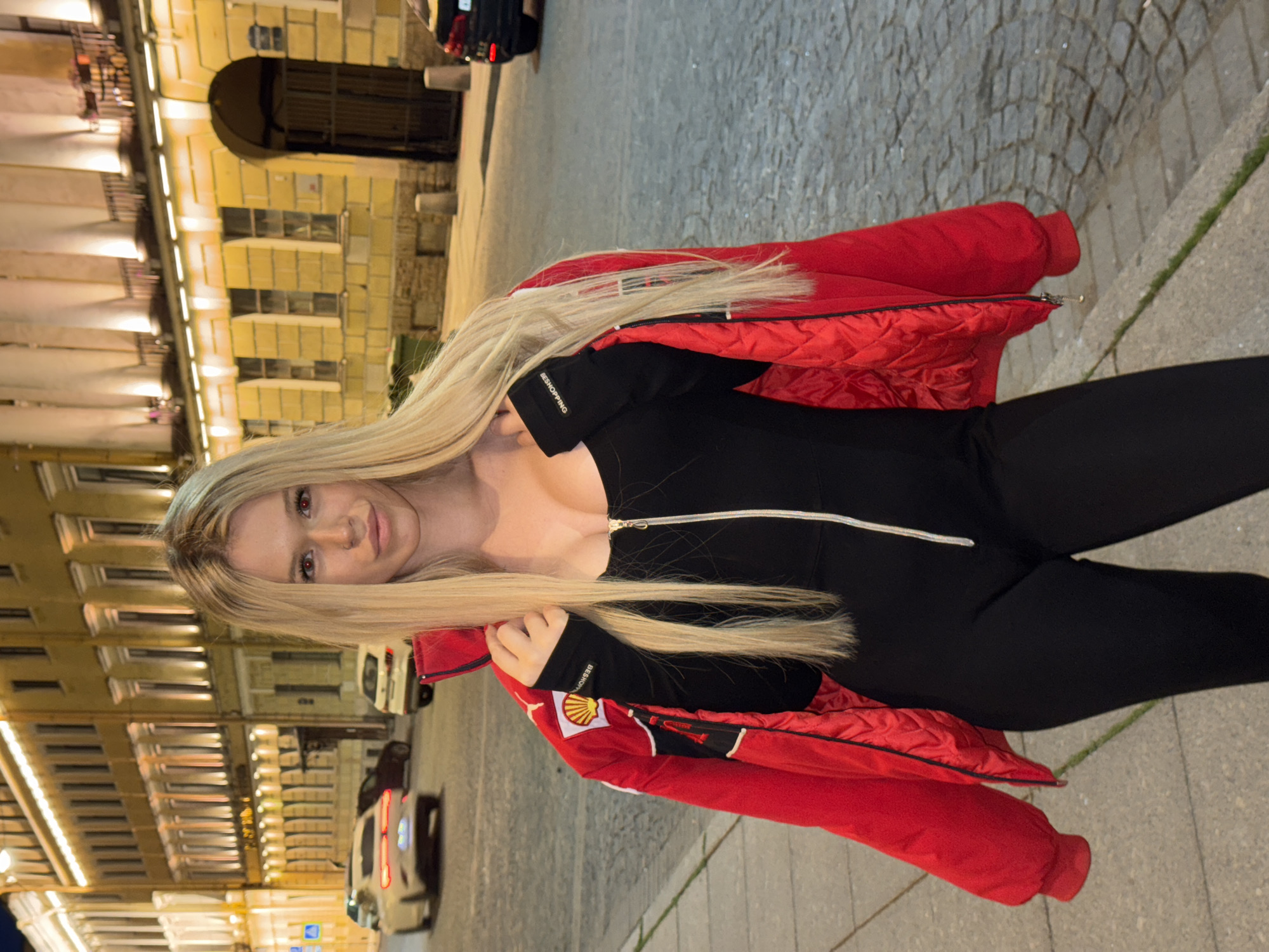 A blonde woman with red-tinted eyes is photographed from the chest up, outdoors at night. She is wearing a black jumpsuit and a red quilted jacket. Her long blonde hair is over her shoulders, and a portion of it is obscuring her face. The jacket has a white zipper and a Shell logo on the left sleeve. The background shows a cobblestone street with buildings on the left. The buildings have illuminated windows and architectural details characteristic of Saint Petersburg, Russia. Cars are visible on the street, and the overall lighting suggests it is nighttime. The woman has a slight smile, and her gaze is directed towards the camera. Text visible on the jacket sleeve reads "BESHOPPING".
