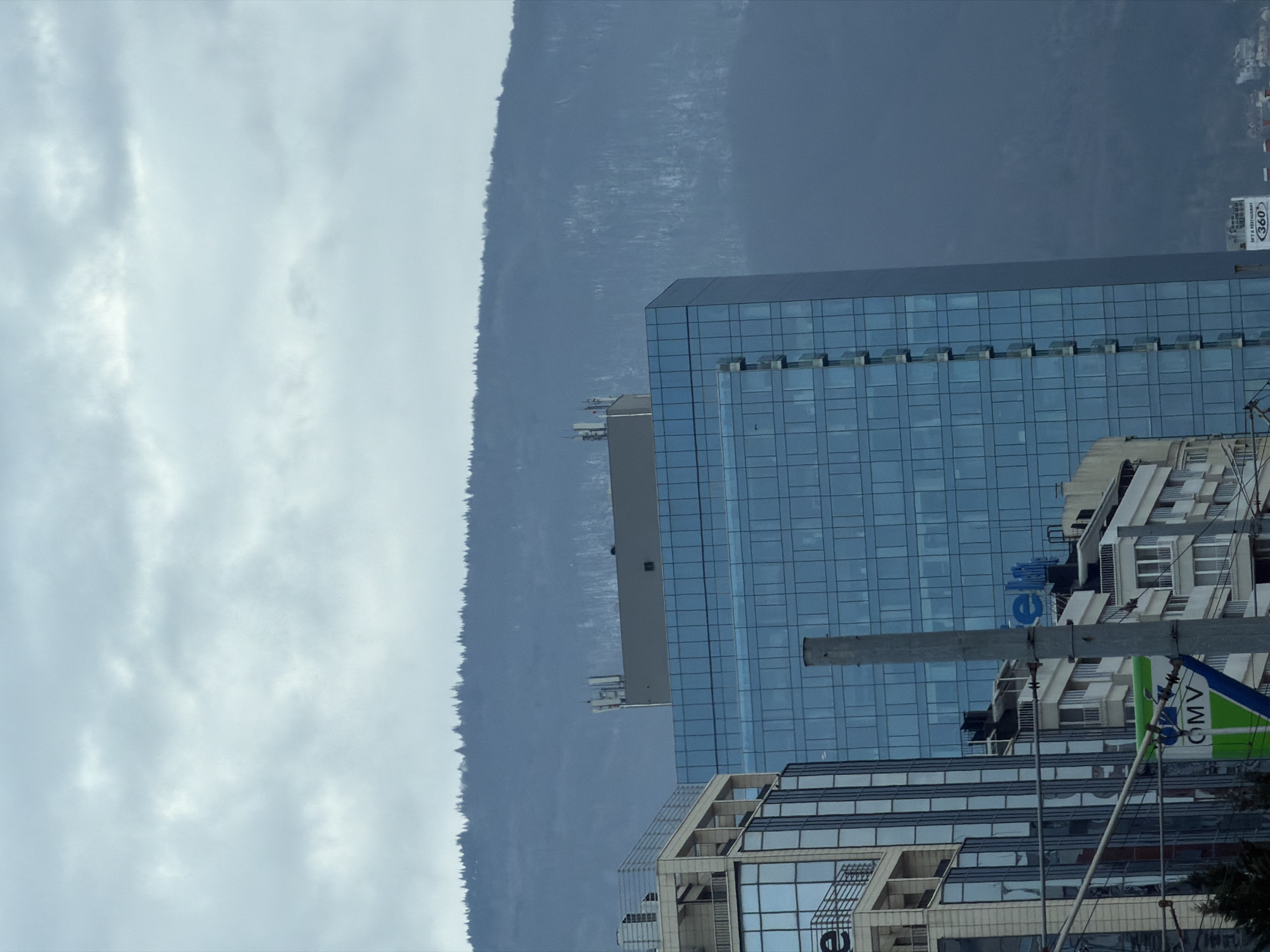 The image shows a view of a city landscape in Sofia, Bulgaria. In the foreground, there is a tall modern glass building, and an older apartment building next to it. Behind the building is a large mountain range covered in trees. The sky is overcast with grey clouds. There are some objects such as antennae on top of the building. The photo is taken on the side, and the city is in the right of the image.