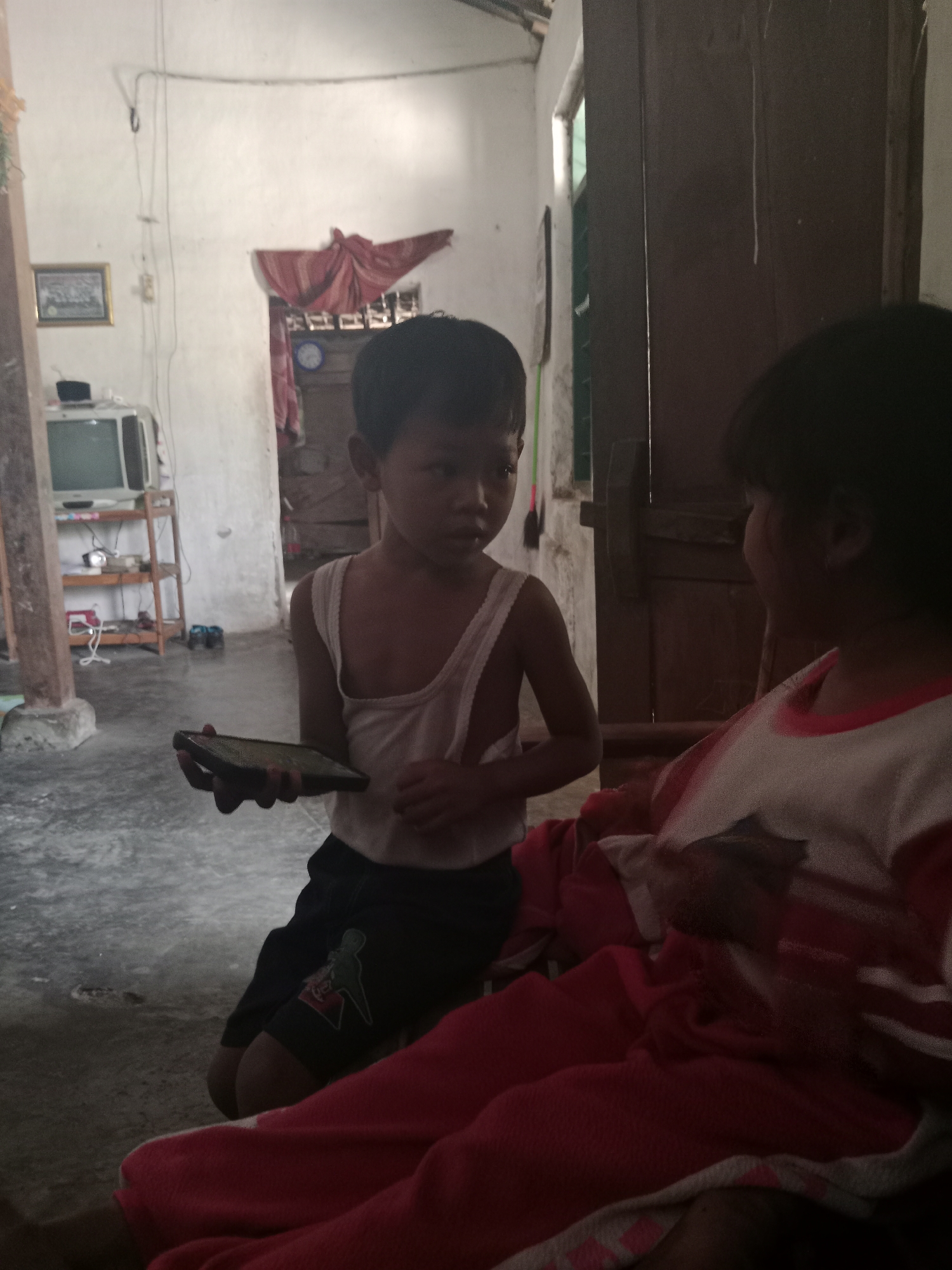 Two children are in a dimly lit room. A boy on the left is kneeling and holding a dark-colored smartphone. He is wearing a white tank top and dark shorts with a graphic design. A second child, whose face is mostly obscured, is to the right, wearing a red and white shirt. The room appears to be a home in Ringinrejo, Indonesia, with a concrete floor and white walls. In the background, a wooden shelf unit holds a television and other items. A red cloth hangs in an open doorway.