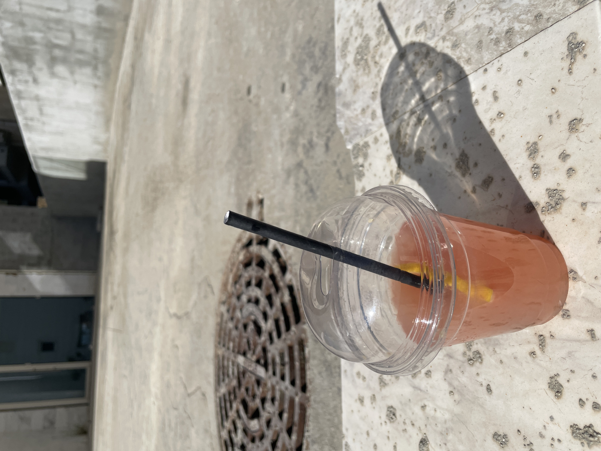 A plastic cup filled with a pink liquid sits on a stone surface, with a black straw sticking out. A shadow of the cup and straw is cast on the stone. To the left, there is a metal grate and a glimpse of a building facade. The location is Boreti, Montenegro.