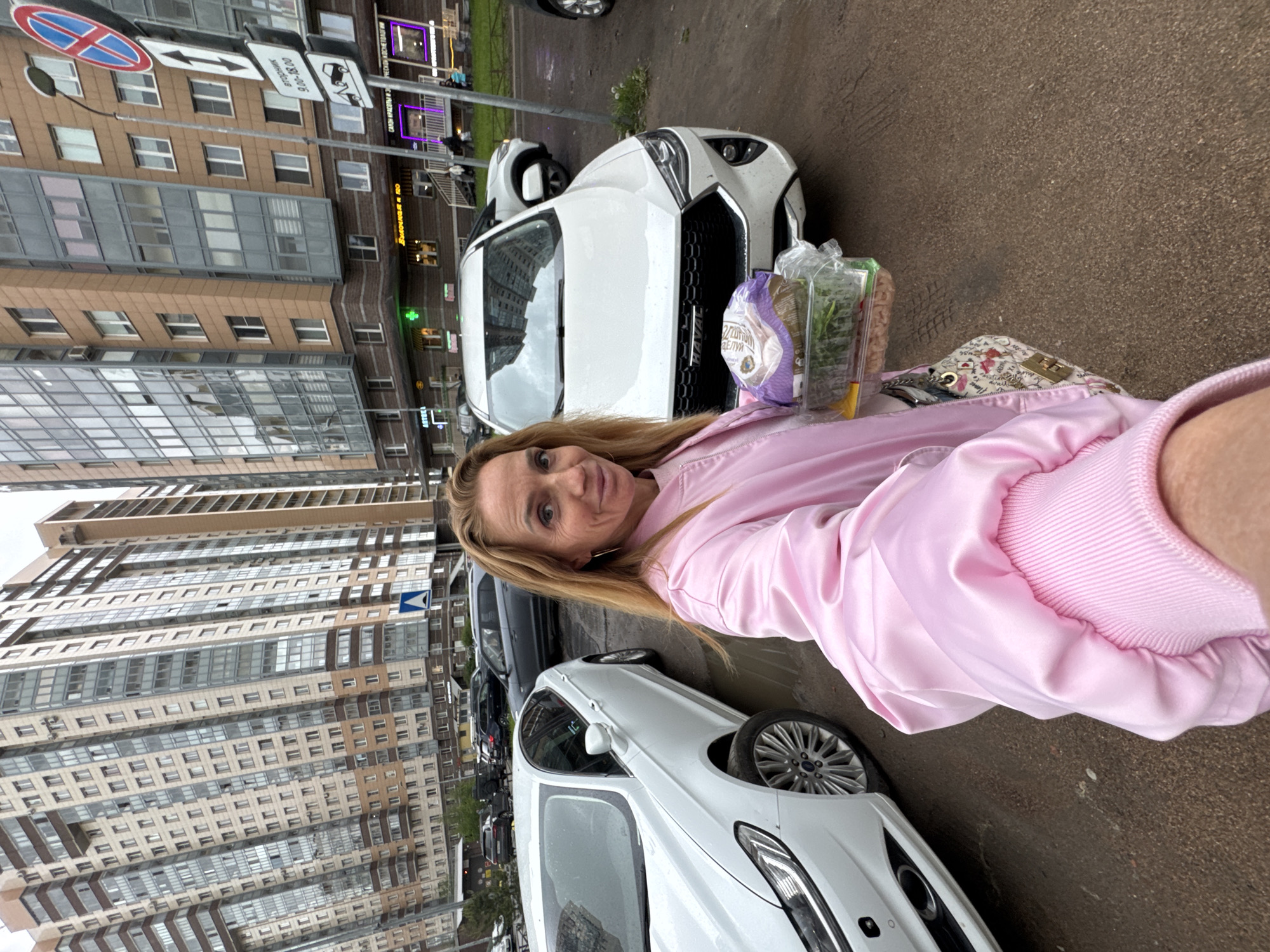 The image shows a woman standing on a city street in Saint Petersburg, Russia. Tall apartment buildings are visible in the background. The woman is holding groceries next to a white car. Street signs are also visible in the background.