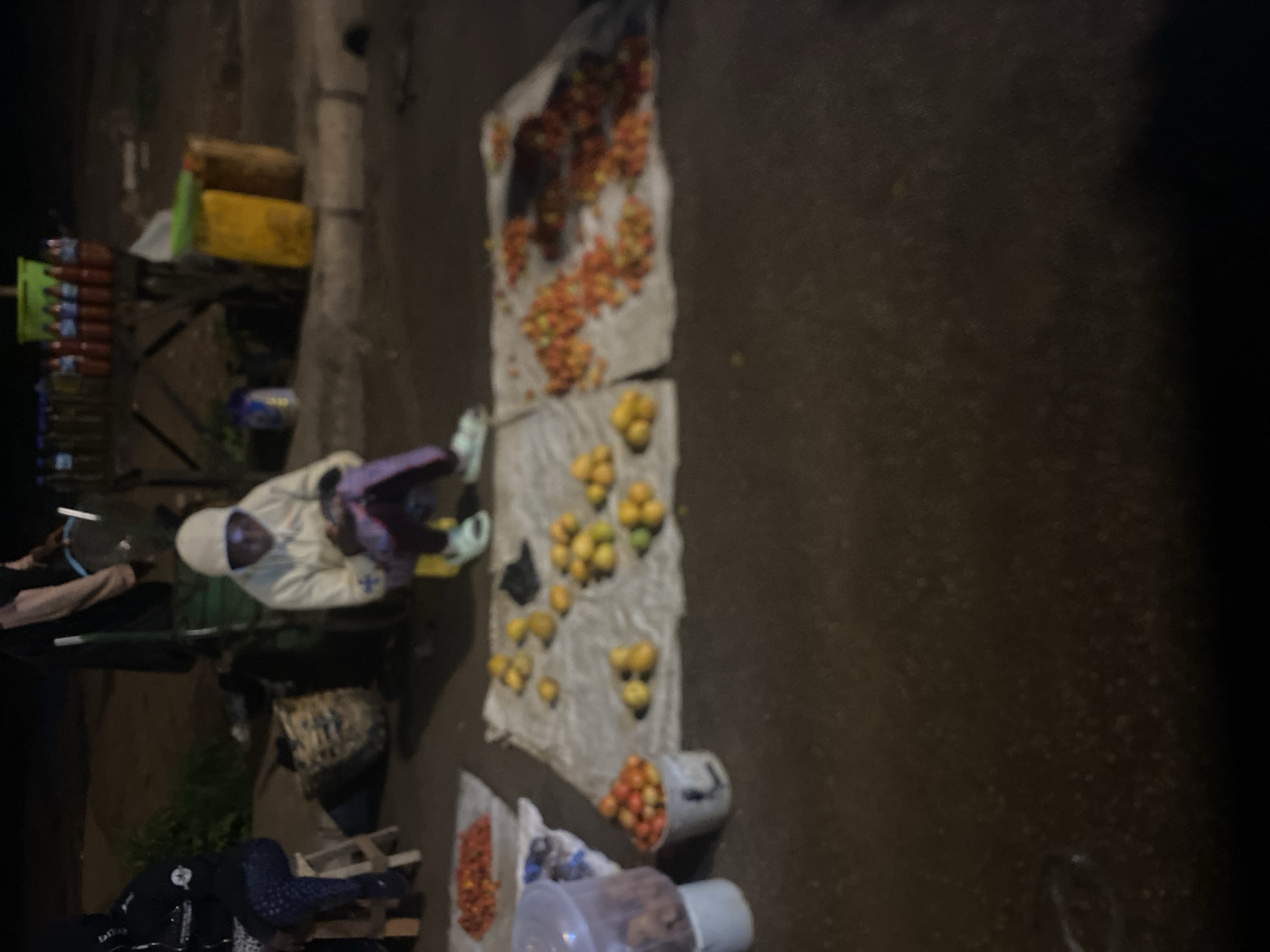 This image, taken from an elevated perspective looking down, captures a nocturnal street market scene in Bukuru, Nigeria. The dim, likely artificial, lighting suggests it is nighttime or very early morning.
On the ground, various types of fresh produce are laid out on light-colored cloths or tarps. In the upper-middle section, a substantial pile of small, reddish-orange fruits, resembling tomatoes or small oranges, is visible. Below this, another spread features yellow, round fruits, likely oranges or tangerines. A smaller collection of similar reddish-orange fruits is positioned towards the bottom right.
To the left of the frame, a makeshift stall or cart holds several clear bottles with red and green labels, stacked vertically, likely containing beverages. Behind these bottles are stacked yellow and brown crates or boxes. Near the bottom right, a clear plastic container with a lid, possibly holding nuts or other snacks, is visible.
Several people are present. One individual, wearing a light-colored hooded top, stands with their back to the camera on the left side of the scene, seemingly attending to the stall. Another person is partially visible in the lower-left corner, wearing dark clothing. The figures suggest vendors or customers engaged in trade.
The ground appears to be a paved or unpaved street or market area, possibly damp in places. The overall atmosphere is that of a bustling, albeit dimly lit, commercial activity. No discernible text is visible in the image due to the distance, low light, and blur.