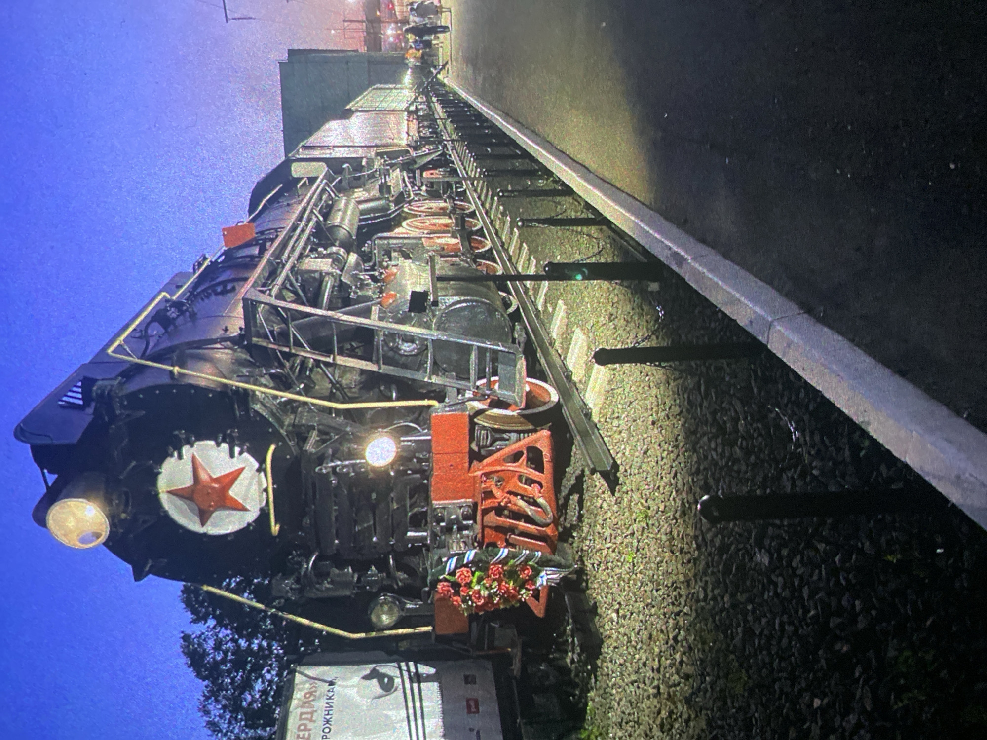 A large, black steam locomotive is prominently displayed on railroad tracks at what appears to be a station or depot, illuminated by artificial lights at night or dusk. The image is oriented vertically, showing the train's front and side.
The locomotive is well-maintained, featuring a classic design with numerous pipes, gauges, and mechanical components visible along its length. Its primary color is black, accented with red on the cowcatcher, some wheel parts, and details on the front. A large, white disc with a prominent red five-pointed star is affixed to the very front of the locomotive, a characteristic emblem of Soviet-era machinery. Below this star, a small wreath of red flowers, interspersed with blue and white ribbons, is placed on the cowcatcher, suggesting a commemorative or ceremonial purpose.
The tracks are visible beneath the train, with gravel ballast illuminated by strong lights. To the right of the tracks, a dark asphalt platform edge runs parallel to the train. The background is dark, with hints of industrial or building structures faintly visible under distant lights. The sky is a deep blue, indicative of night.
On a white sign at the bottom left of the original image (front right of the train in the image), partial Cyrillic text is visible, reading "ЕРДЯ" and "рожника", further confirming the Russian context of Saint Petersburg. The artificial lighting creates strong contrasts, highlighting the train's features against the dark surroundings. No people are visible.