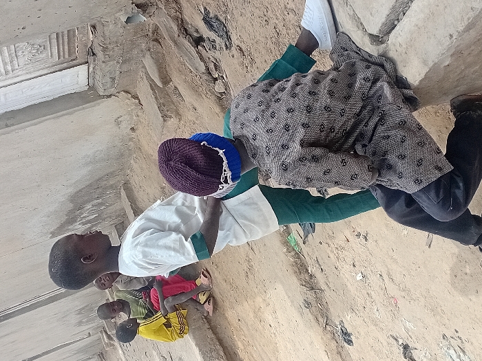 The scene depicts five young males congregating outdoors in the city of Ayama, Nigeria. In the foreground, an individual sits on a concrete ledge with their back to the viewer, wearing a dark purple and blue knitted beanie, a grey patterned long-sleeved shirt, and dark trousers. Their right foot, clad in a white sneaker, rests on the ledge. Adjacent to them, another male stands, facing right, dressed in a white long-sleeved top with green cuffs and green trousers. Further in the background to the left, three other males are seated on a lower concrete ledge. One wears a yellow top and shorts with pink sandals, another a green and black striped long-sleeved top, and the third a light brown long-sleeved top. The ground is unpaved dirt with scattered debris, including dark plastic. Unpainted concrete building walls form the background, with a decorative light-colored wooden door visible on the right. The overall environment appears to be an urban or semi-urban outdoor area.
