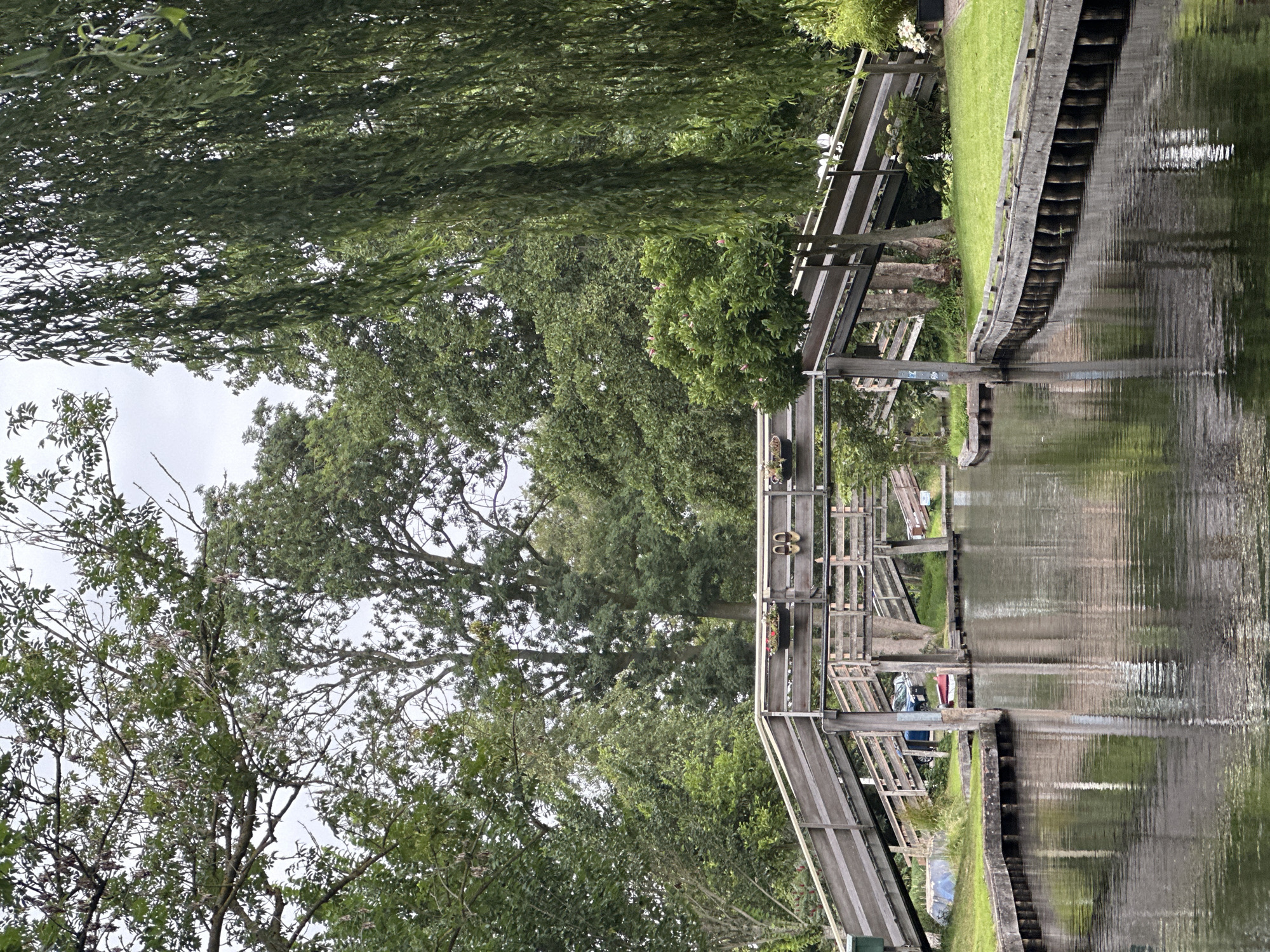 The image depicts a serene canal scene in Giethoorn, Netherlands, a village renowned for its waterways and lack of roads. A wooden bridge, with its distinctive ramped approach, spans the dark, reflective water. The bridge is adorned with flower baskets, and on its railing, a pair of flip-flops are casually placed. On the left bank, a blue boat and a red canoe are visible near the grassy edge, suggesting a peaceful, recreational environment. Lush green trees, including a weeping willow on the right, frame the scene, their foliage creating a dappled canopy overhead. The water's surface mirrors the bridge and the surrounding greenery, creating a tranquil, almost painterly effect. The overcast sky lends a soft, diffused light to the scene, contributing to the overall sense of calm.