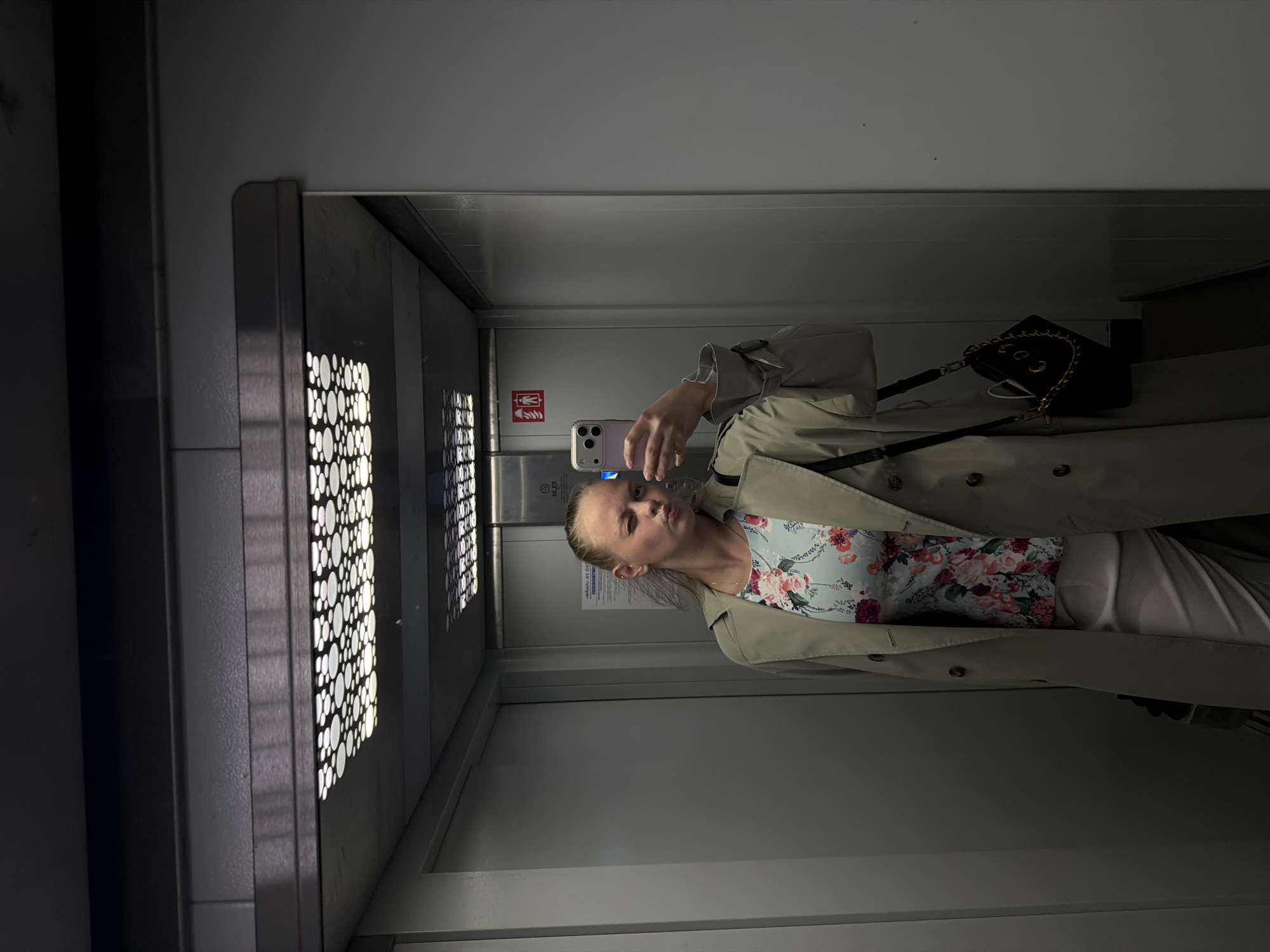 A woman is taking a selfie in an elevator. She is wearing a trench coat over a floral dress and has a black purse hanging from her shoulder. The elevator has a perforated metal wall with a grid of circular cutouts and a sign with a red pictogram. The elevator appears to be in Izhevsk, Russia, based on the assumption that this is the location.