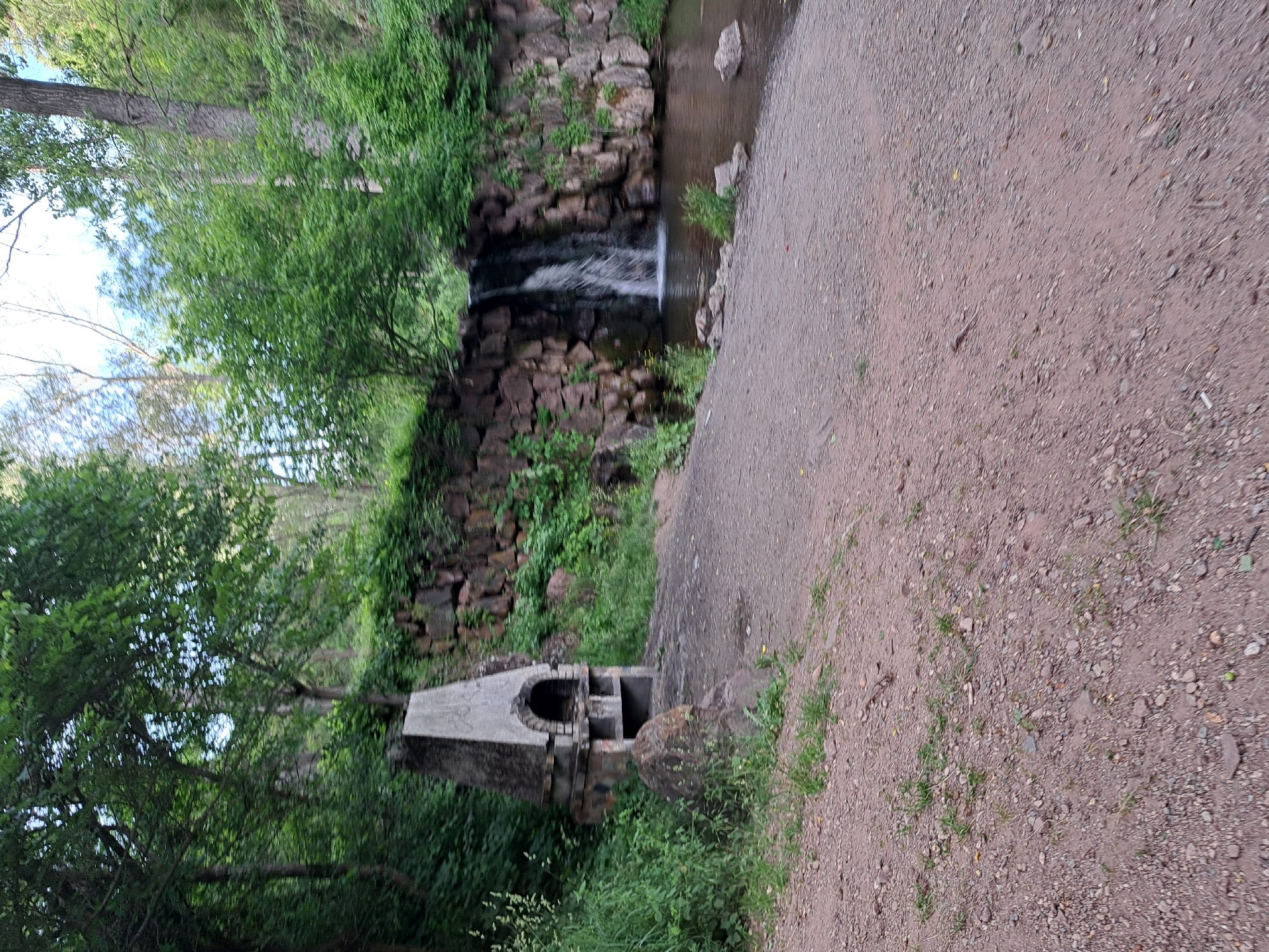 A vertical, slightly blurry image depicts a natural outdoor scene featuring a small waterfall cascading over a stone wall into a narrow stream. To the right of the stream, a gravel path runs parallel to it. In the foreground, to the left, is a rustic, stone barbecue pit with an arched opening. Lush green trees and foliage surround the area, suggesting a wooded or park-like setting. The lighting implies it is daytime, possibly overcast or with dappled sunlight filtering through the leaves. The scene evokes a sense of tranquility and natural beauty. No people are visible, and there is no visible text in the image. The location is identified as Lokorsko, Bulgaria.